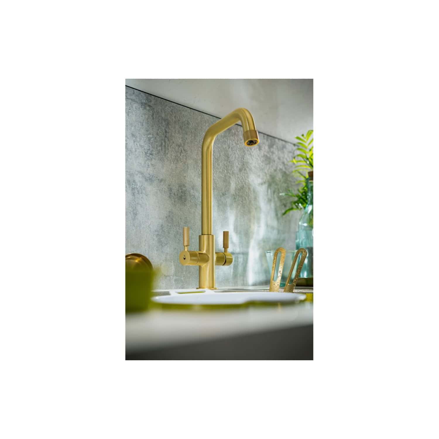 Brass kitchen tap with a modern design, mounted on a kitchen counter with a textured grey backsplash, featuring minimalist fixtures and a glass vase with green foliage for a contemporary bathroom aesthetic.