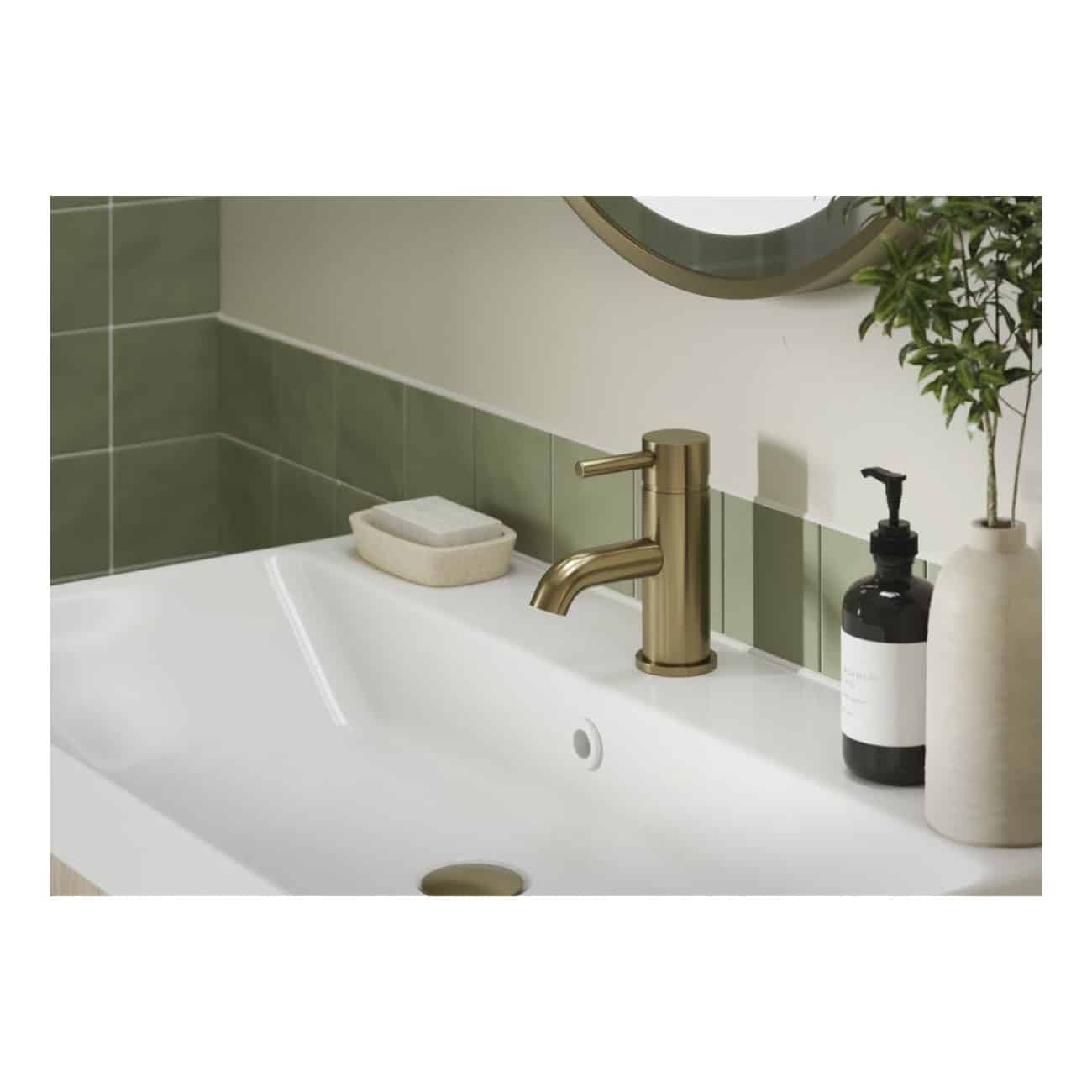 Sleek white bathroom sink with a modern brass faucet, green tiled backsplash, and decorative elements including a soap dispenser and a potted plant, creating a stylish and functional space.