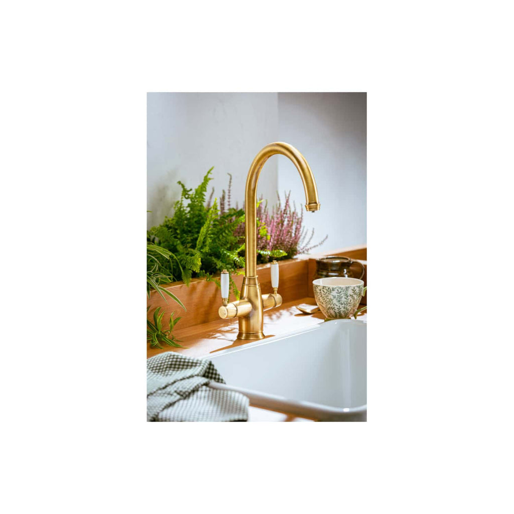 Polished gold kitchen sink faucet with vintage handles, set against a wooden countertop with potted herbs and a patterned ceramic cup, creating a stylish and functional kitchen detail.