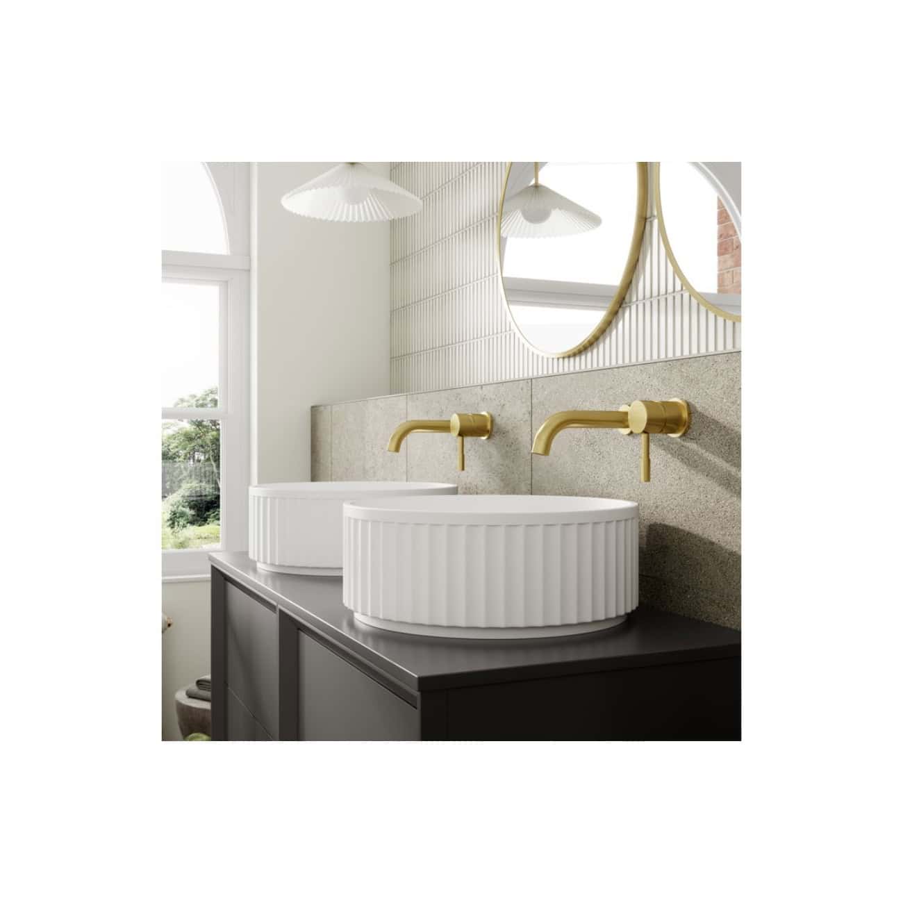 Sleek modern bathroom featuring white oval vessel sinks with brass wall-mounted faucets, a large round mirror, and natural light from a window, creating a contemporary and stylish space.
