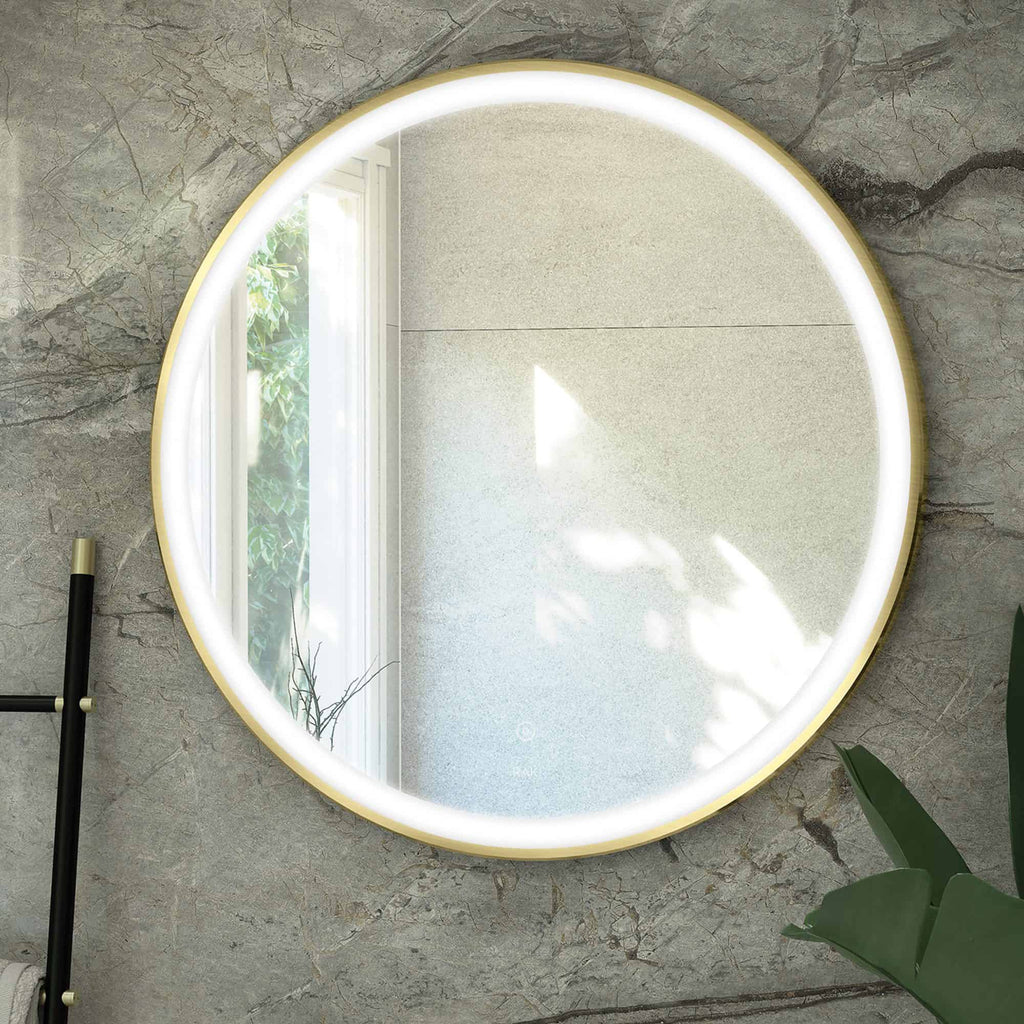 Round LED illuminated bathroom mirror with a gold trim, mounted on a textured grey wall, reflecting a window with natural light and greenery, ideal for modern bathroom decor.