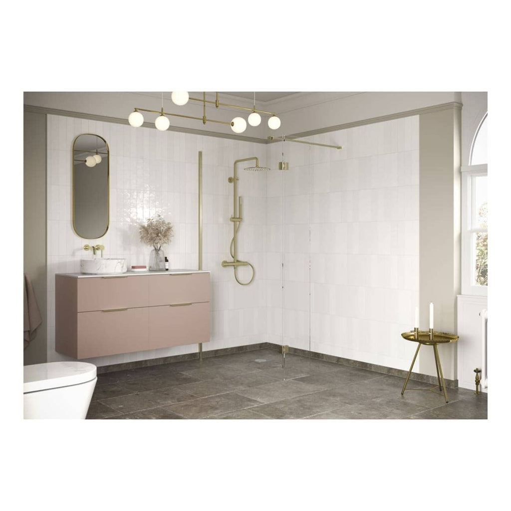 Modern minimalist bathroom with light pink floating vanity, gold fixtures, walk-in shower with glass partition, white tiled walls, grey floor tiles, and elegant decor including a mirror, dried flowers, and a small side table.