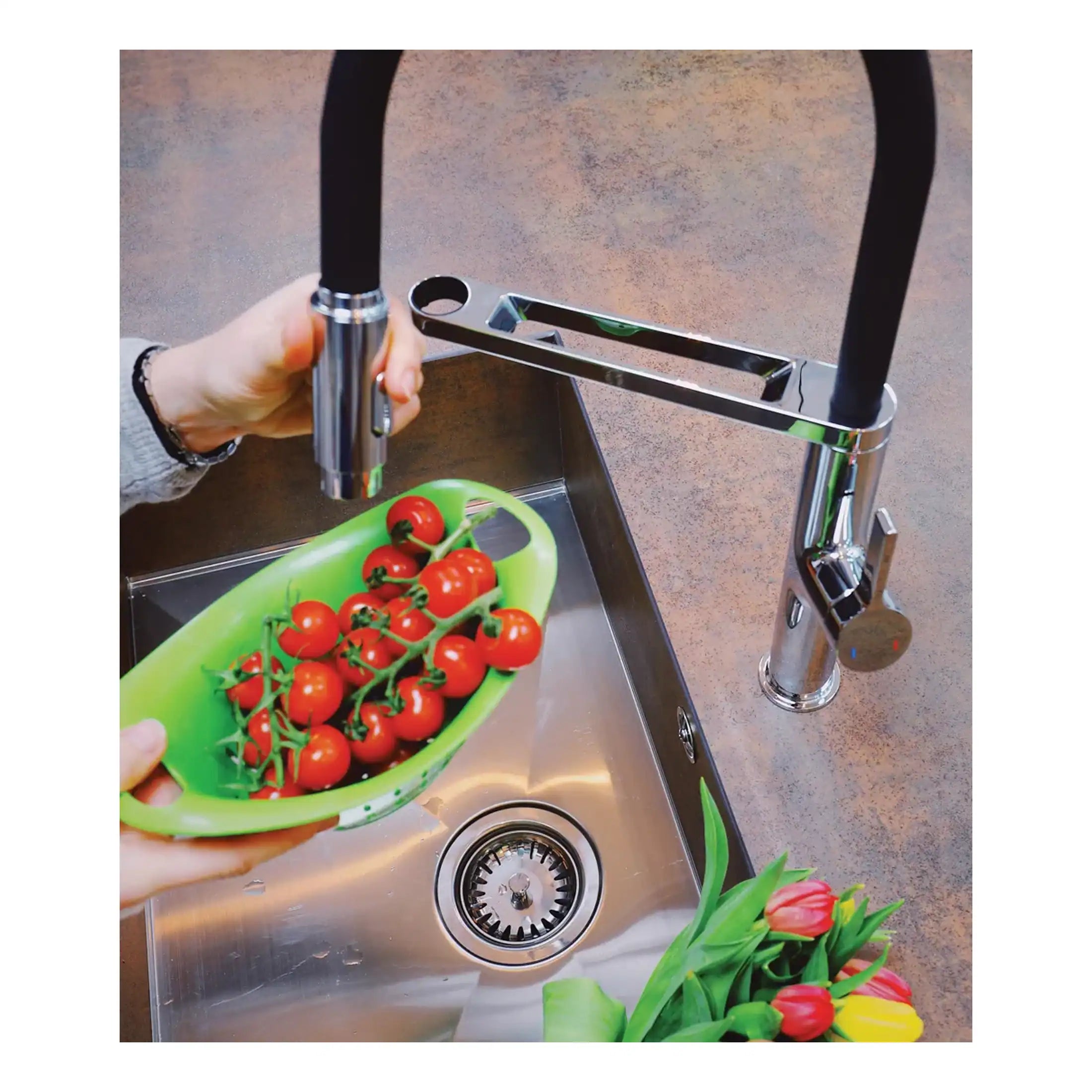 Bright modern kitchen stainless steel single basin sink with chrome finish, high-arc kitchen tap, and a hand holding a green bowl of cherry tomatoes, surrounded by fresh tulips, showcasing stylish kitchen spaces.