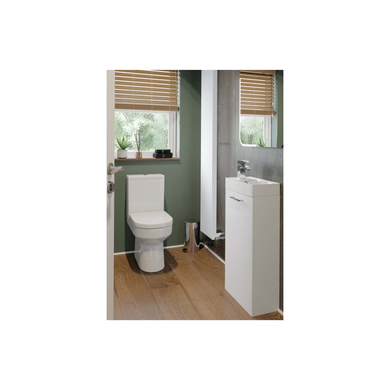 Modern compact bathroom with a white toilet, small white sink cabinet, wood flooring, and green walls, featuring natural light from a window with wooden blinds, designed for efficient space utilisation.