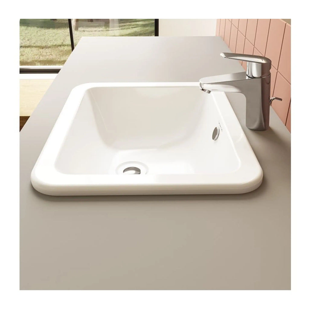 White ceramic bathroom sink with modern chrome faucet on a beige countertop, with pink tiled wall and window in the background.