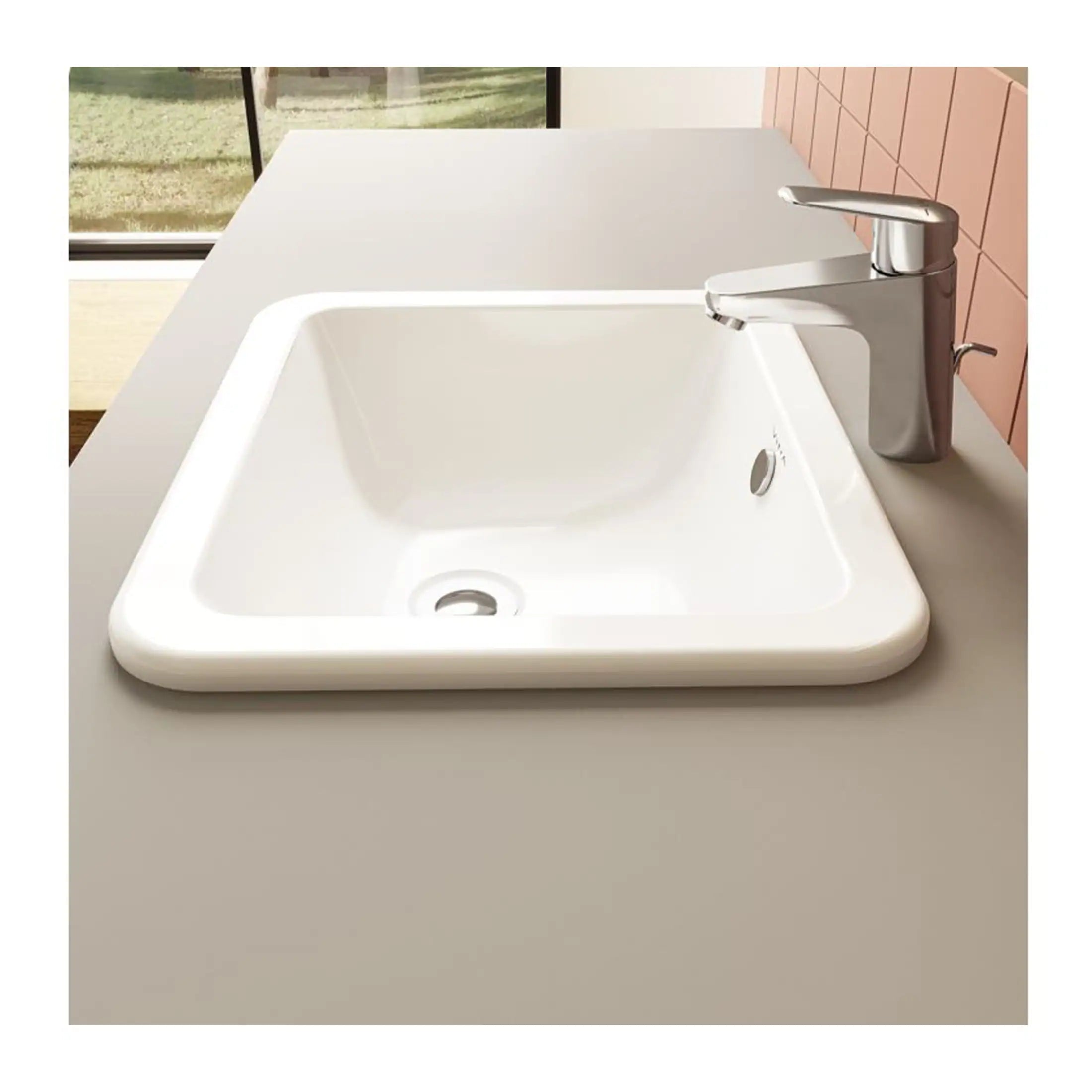 White ceramic bathroom sink with modern chrome faucet on a beige countertop, with pink tiled wall and window in the background.