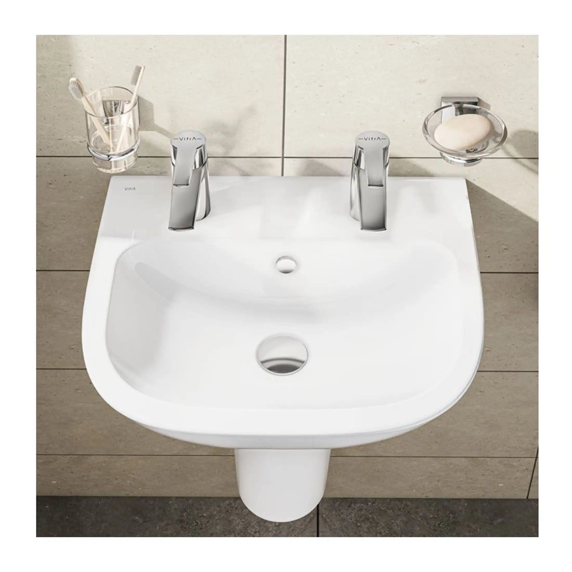 Shiny white bathroom washbasin with dual chrome taps, toothbrush holder, soap dish, and beige tiled wall background, showcasing modern bathroom fixtures and design.
