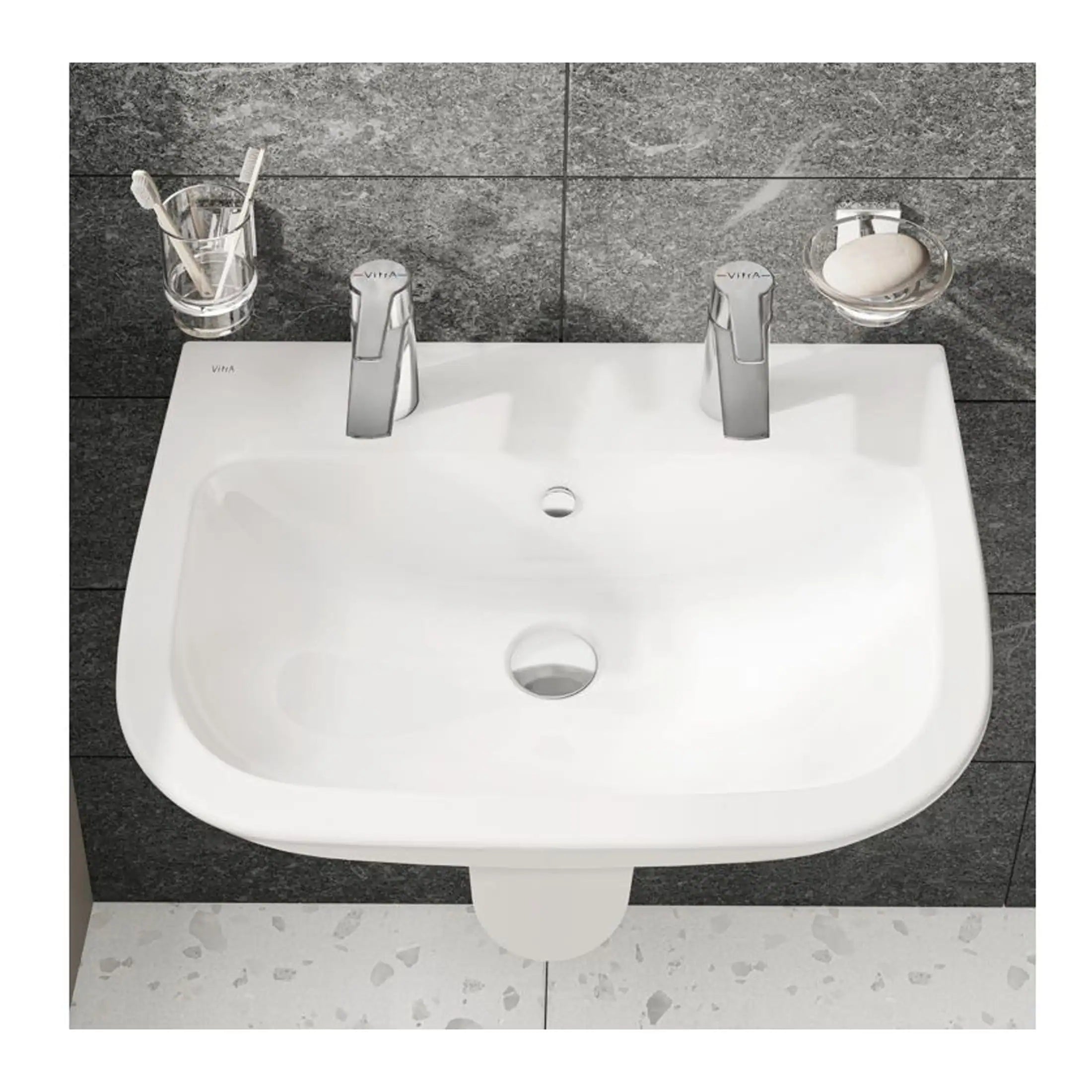 Shiny white bathroom sink with dual modern chrome taps, mounted on a dark tiled wall, with accessories like toothbrush holder and soap dish for contemporary bathroom decor.