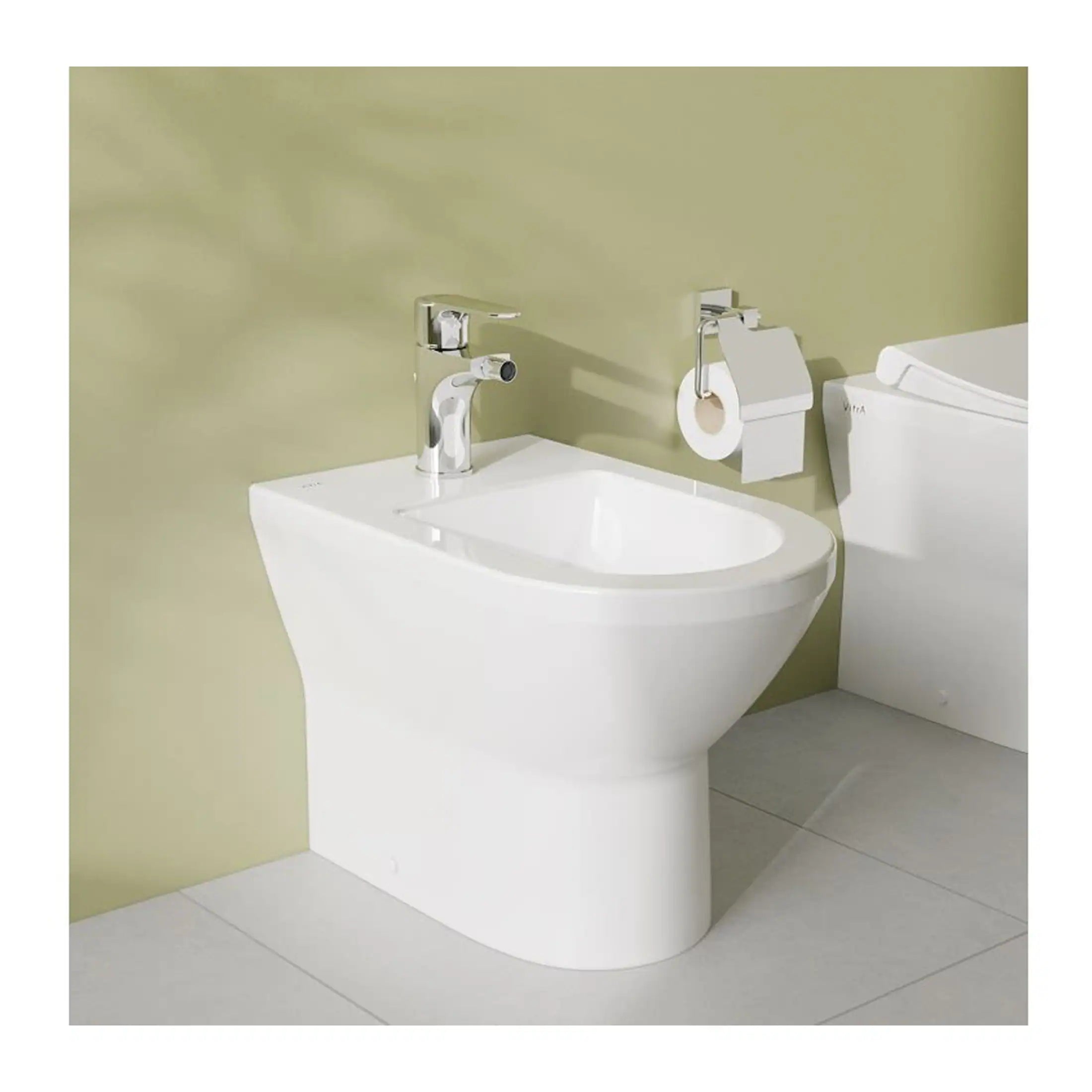 Bidet bathroom fixture with modern chrome faucet and adjacent toilet with toilet paper holder, installed on a light-coloured tiled floor in a contemporary bathroom setting.