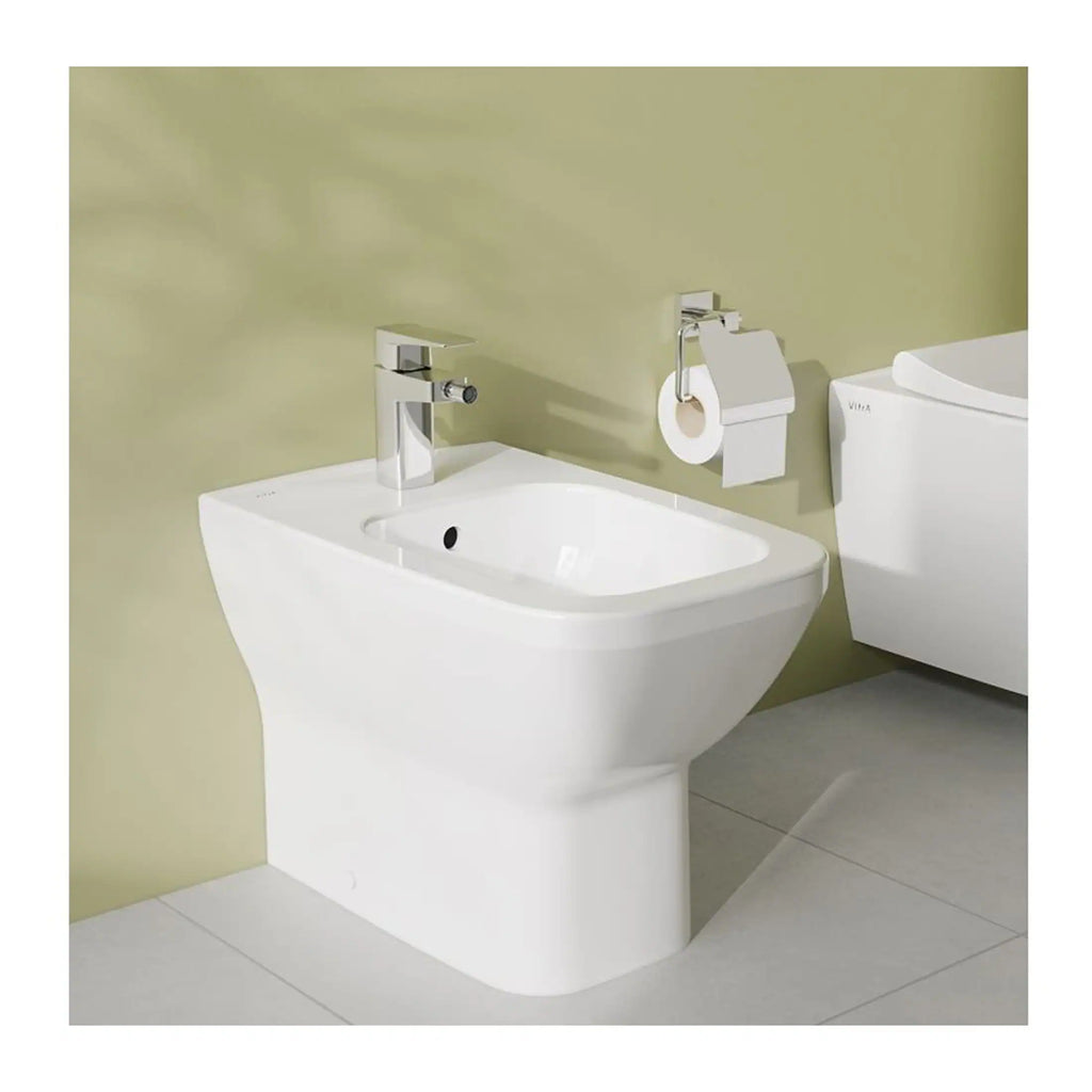 A modern white bidet with a sleek chrome faucet, positioned next to a toilet with a toilet paper holder on a light green bathroom wall, creating a contemporary and functional bathroom setup.