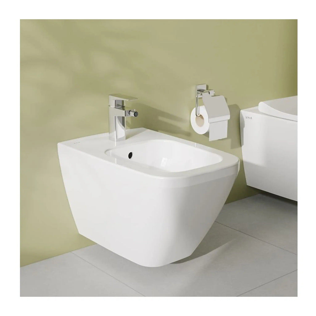 Minimalist wall-mounted bathroom bidet with modern chrome faucet, adjacent toilet with soft-close lid, and contemporary toilet paper holder in a sleek, clean bathroom setting.