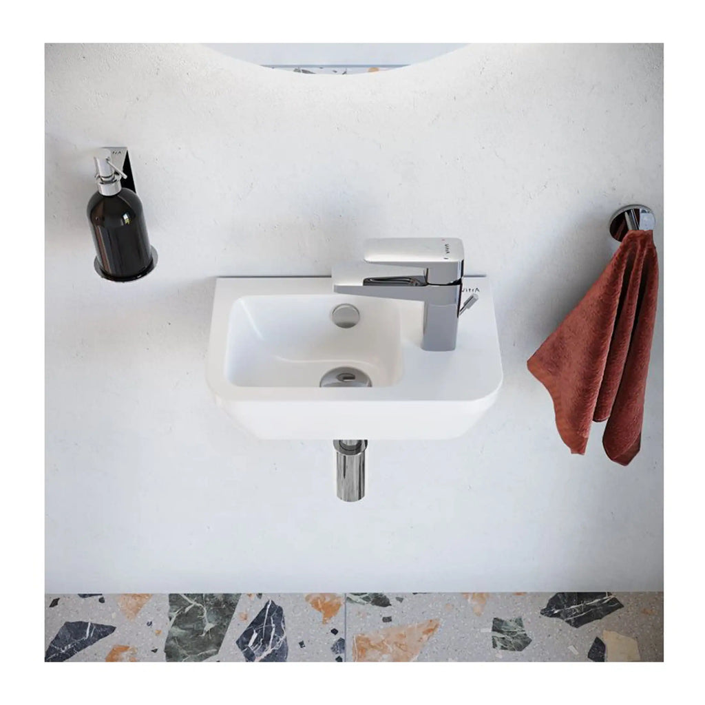 Small white bathroom sink with modern chrome faucet, mounted on wall with a black soap dispenser and rust-coloured towel hanging on the side, set against a plain white wall and terrazzo flooring.