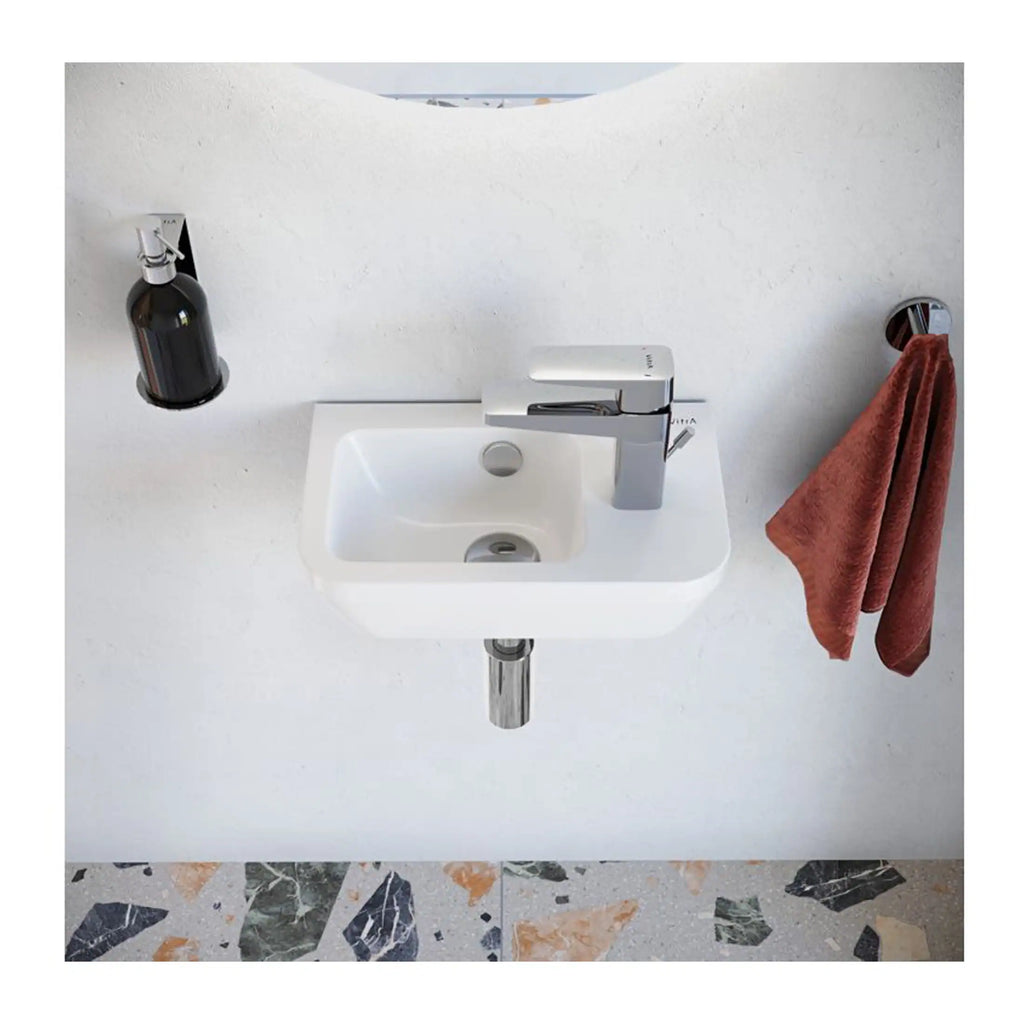 Modern white bathroom sink with chrome faucet, wall-mounted soap dispenser, and rust-coloured hand towel on chrome holder, set against a white textured wall with terrazzo floor, showcasing stylish bathroom design.