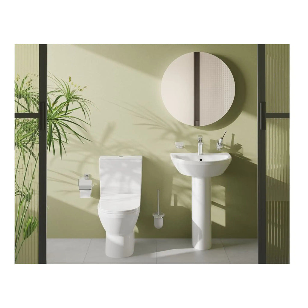 Contemporary white toilet and pedestal sink in a modern bathroom with light green wall, round mirror, and natural sunlight creating shadow patterns. Ideal for bathroom renovation or design ideas.