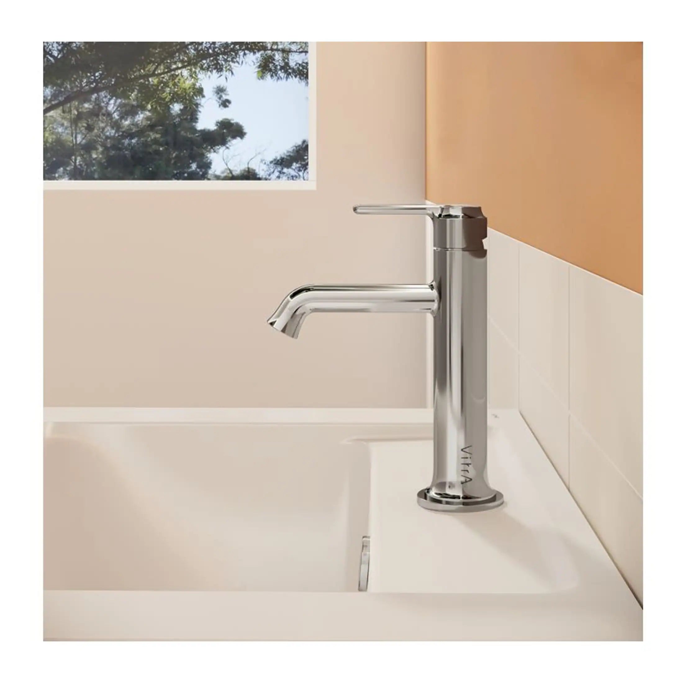 Sleek chrome bathroom tap installed above a white sink in a modern bathroom with beige walls and a window showing trees outside.