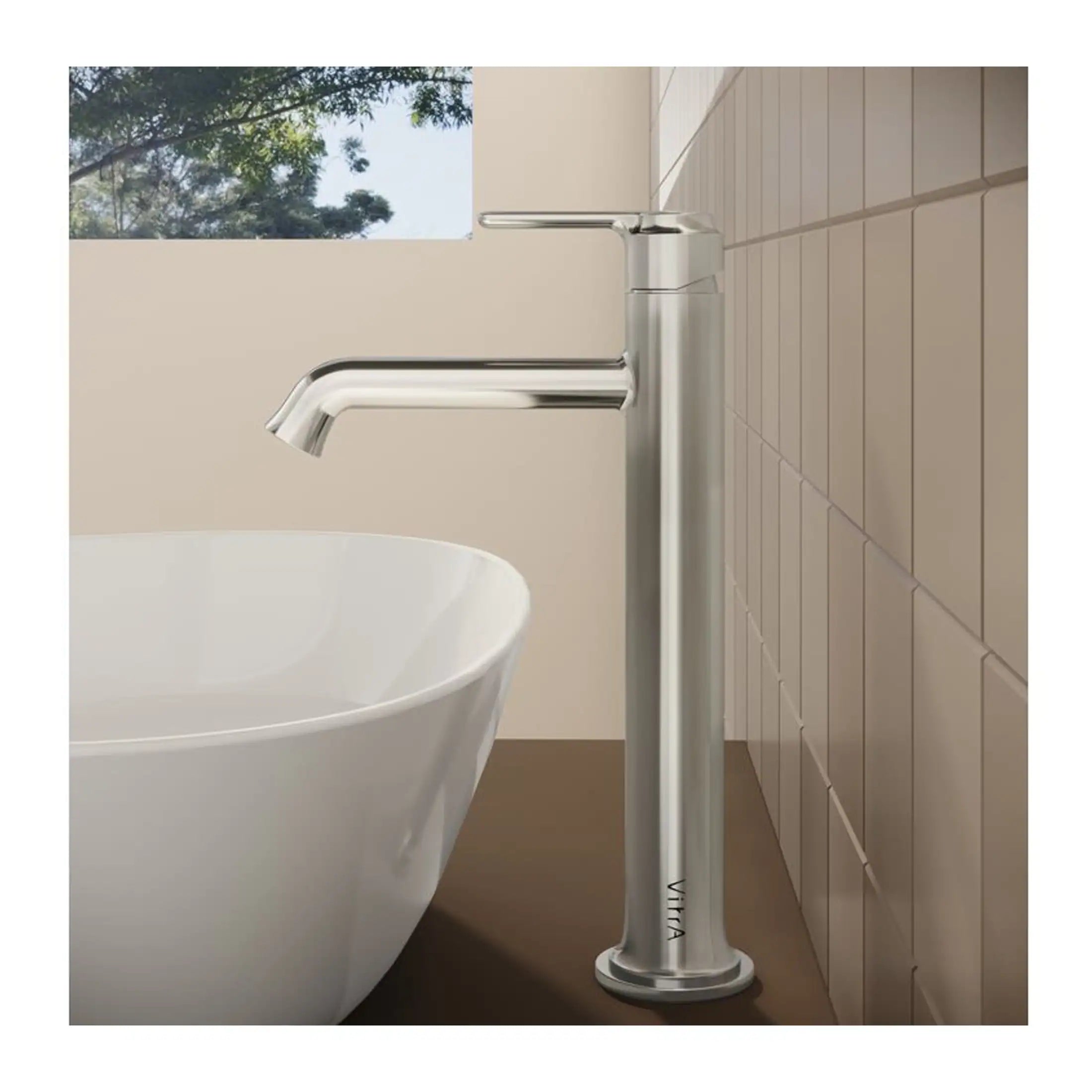 Polished chrome freestanding bathroom tap next to a white bath in a modern bathroom with beige tiles and a window with trees outside.