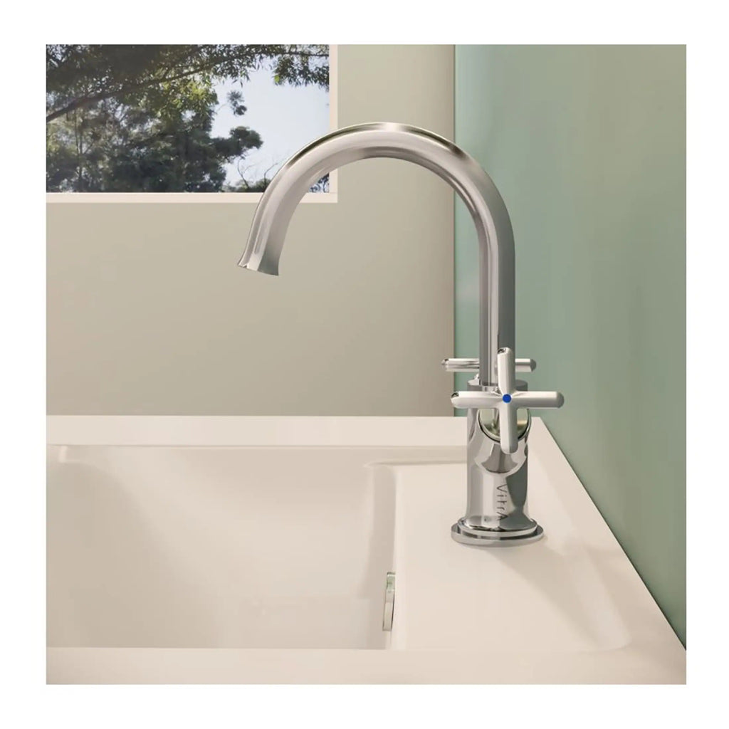 Sleek chrome kitchen or bathroom faucet with a curved spout and cross handles on a white sink with a neutral wall background and a window showing outdoor greenery.