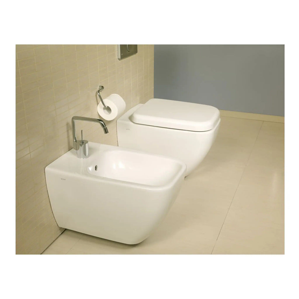 Modern white bidet and toilet in contemporary bathroom setting, featuring sleek design, wall-mounted tap, with beige tiled wall and light flooring, highlighting bathroom fixtures and sanitary ware.