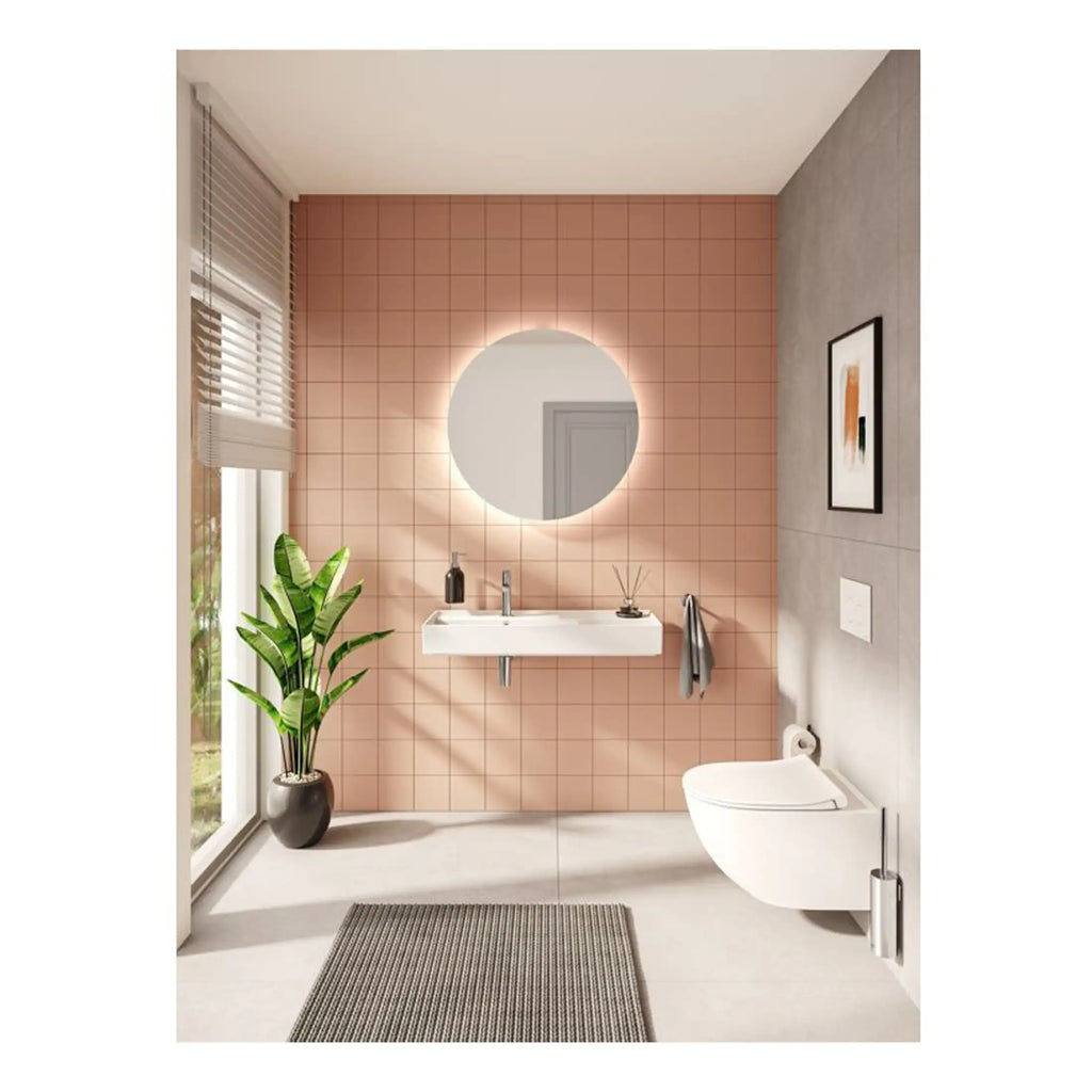 Sleek modern bathroom with pink tiled feature wall, round illuminated mirror, wall-mounted toilet, minimalist sink with accessories, indoor plant, and natural light.