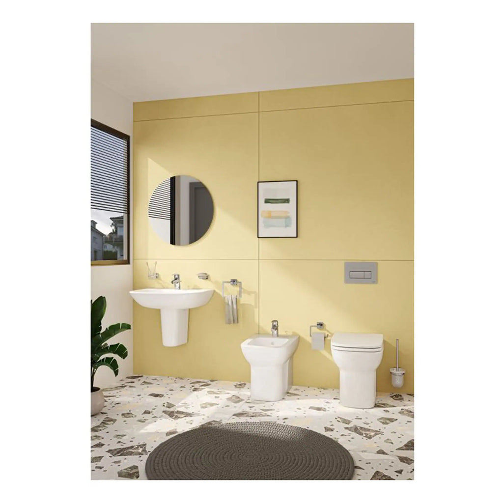 Elegant bathroom with modern white fixtures, including a wall-mounted sink, toilet, and bidet, set against a soft yellow tiled wall, complemented by a round mirror, framed artwork, and a terrazzo floor with a dark rug.