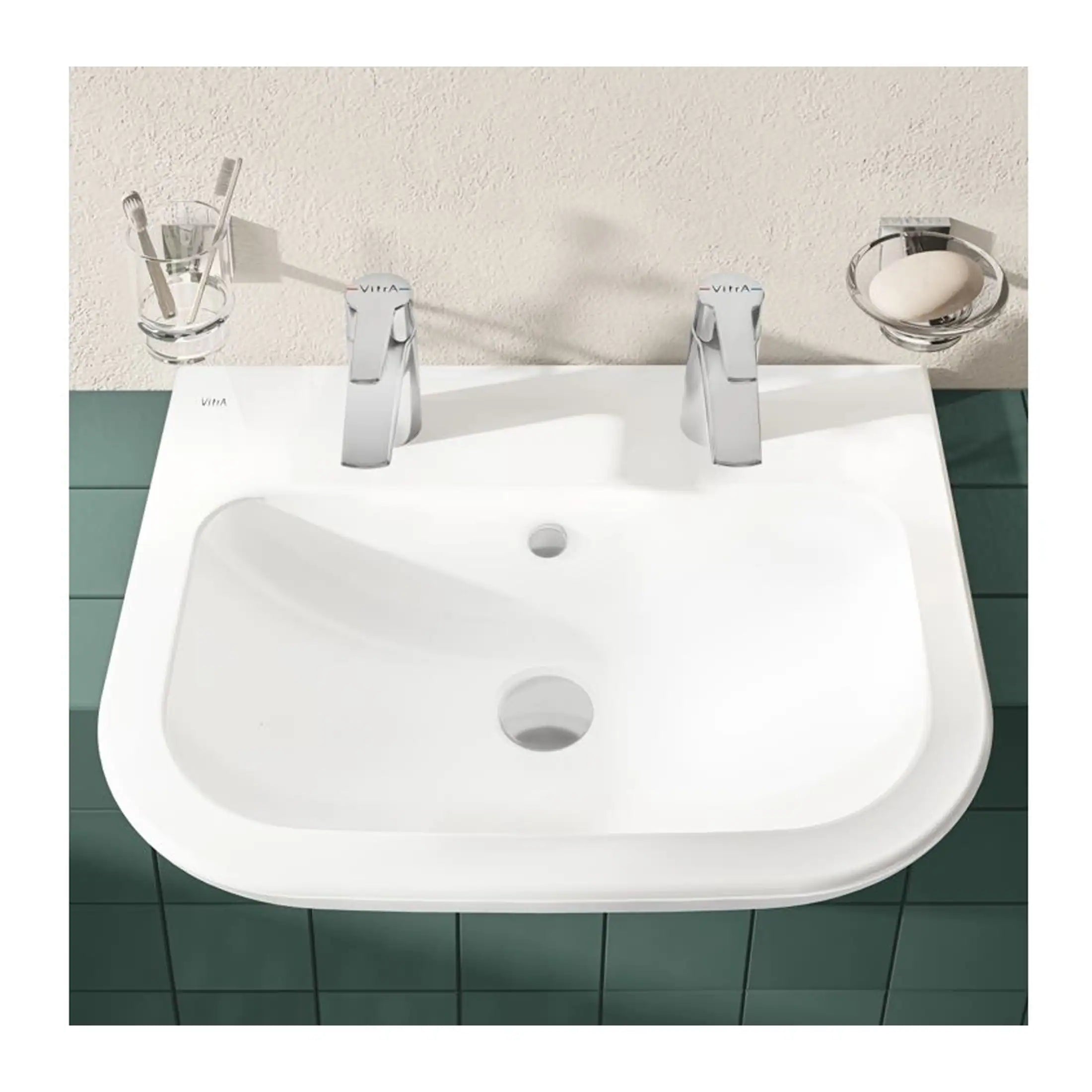 Shiny white bathroom sink with dual modern chrome taps, toothbrush holder with toothbrushes, soap dish with a bar of soap, mounted on green tiled wall for contemporary bathroom decor.