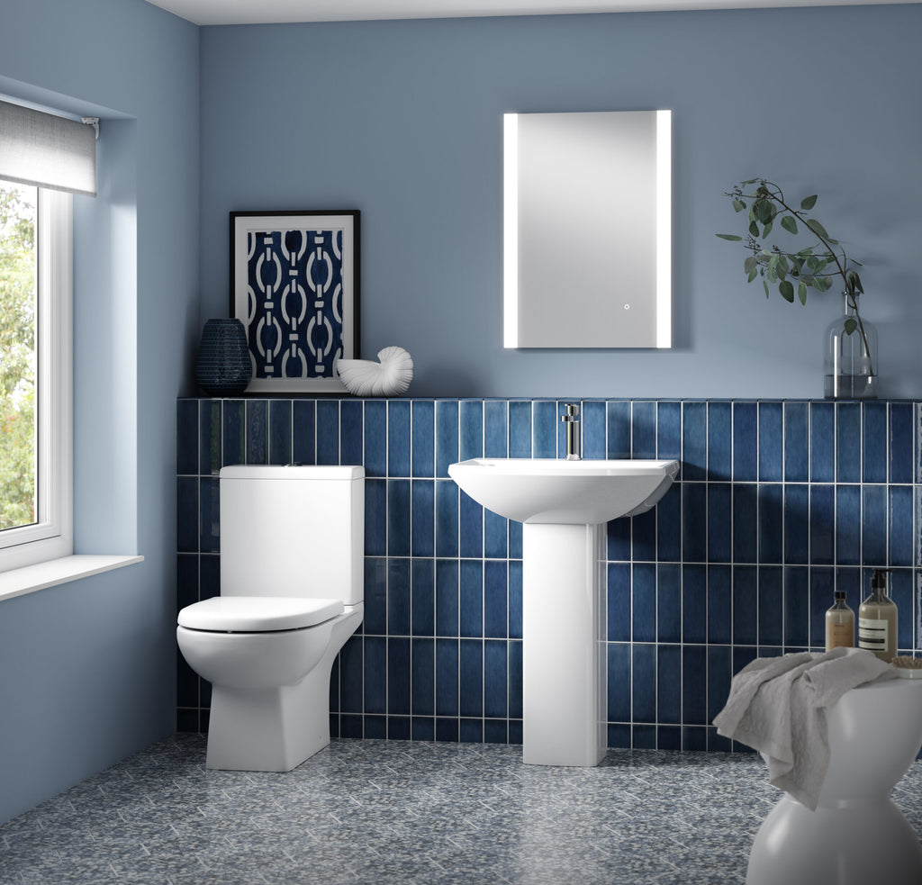 Modern blue tile bathroom with white toilet, pedestal sink, LED mirror, decorative artwork, and natural light, featuring stylish and functional bathroom fixtures and accessories.