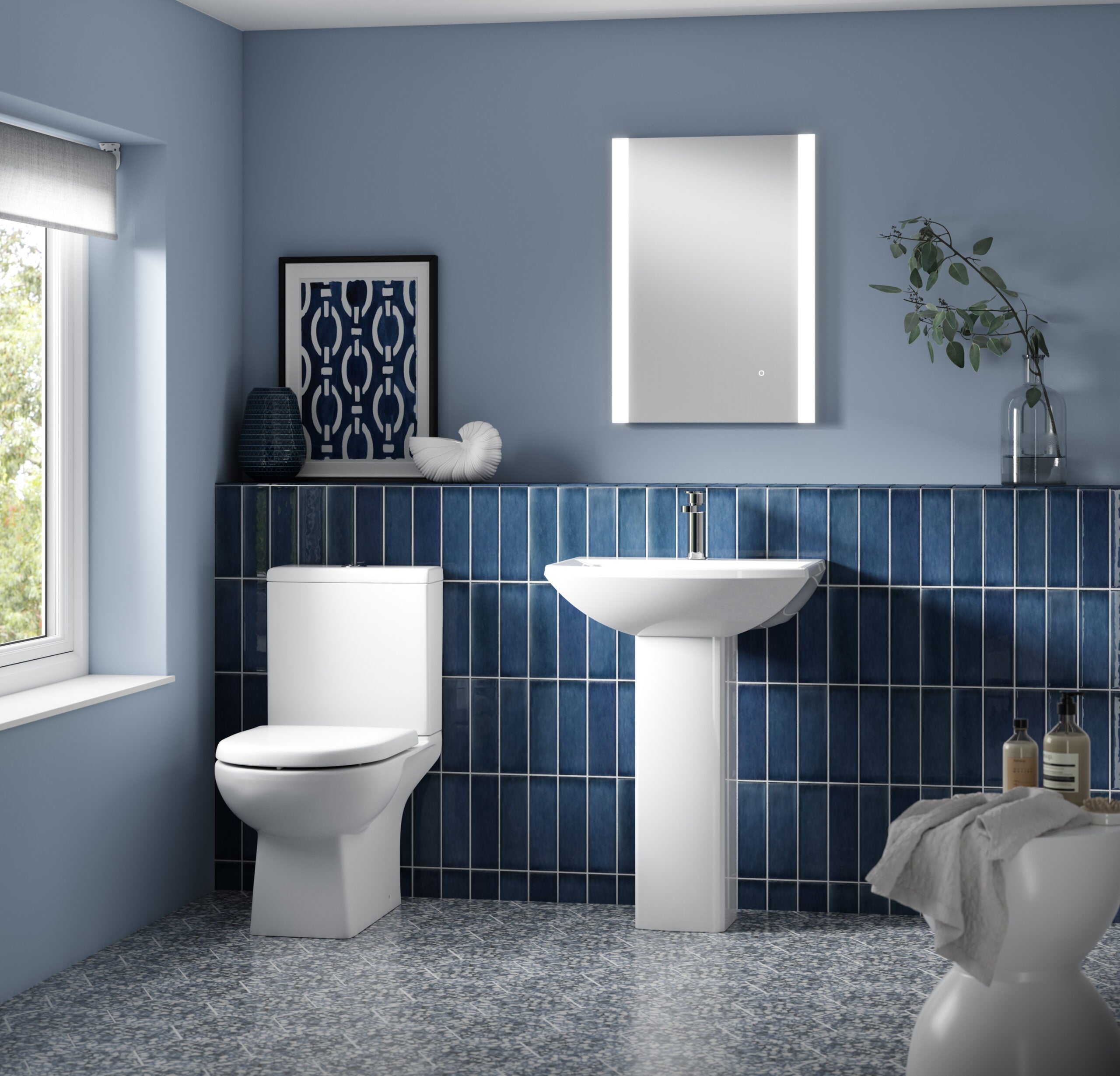 Modern blue tile bathroom with white toilet, pedestal sink, LED mirror, decorative artwork, and natural light, featuring stylish and functional bathroom fixtures and accessories.