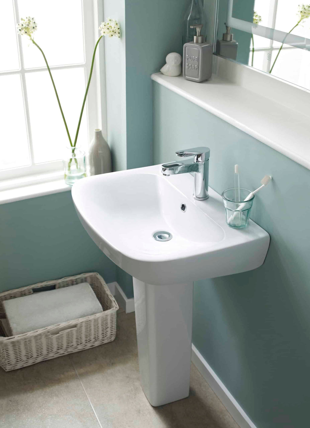 Modern white bathroom washbasin with chrome tap and accessories, situated near a window with decorative flower vase, ideal for contemporary bathroom designs.