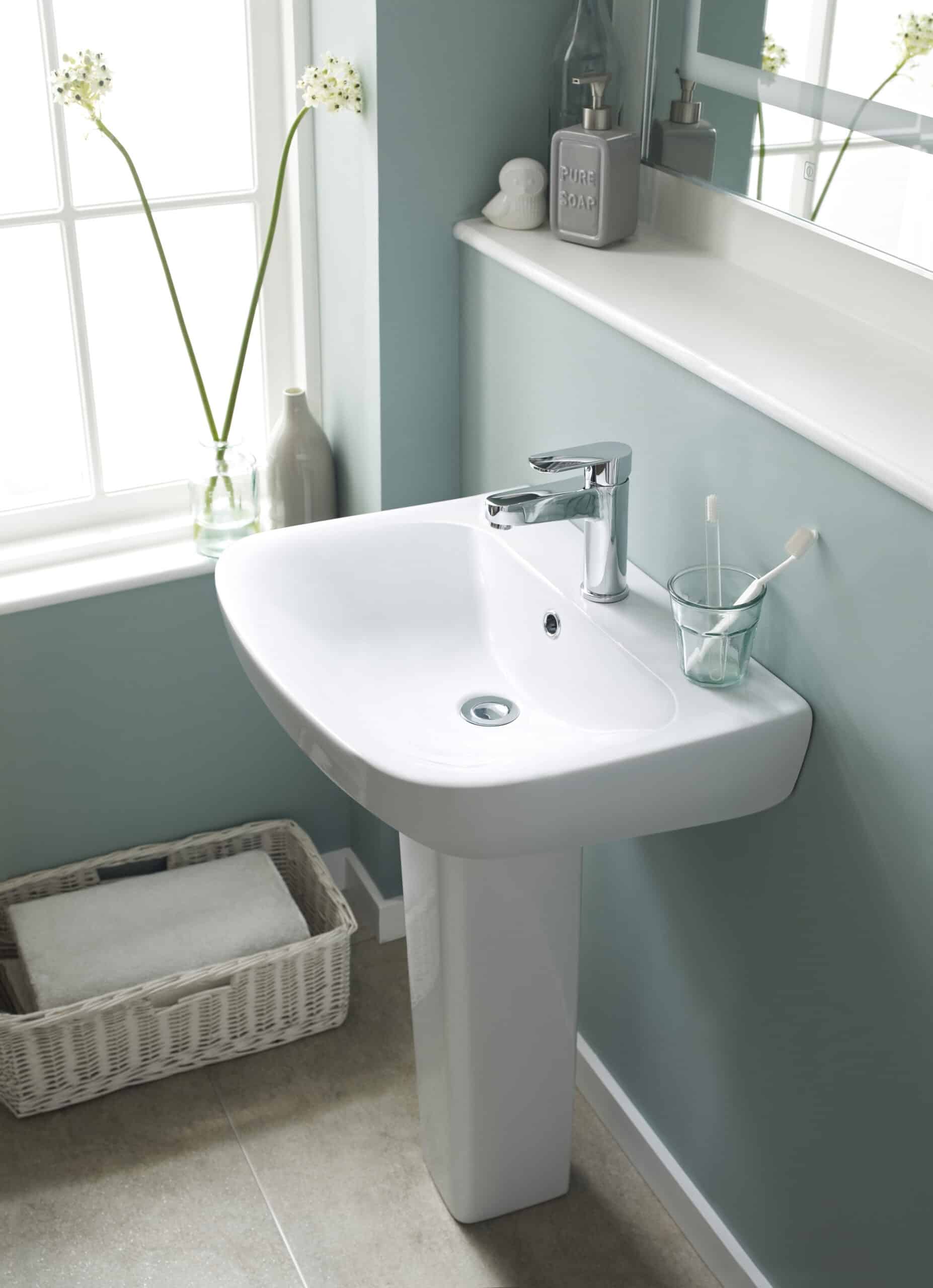 Modern white bathroom washbasin with chrome tap and accessories, situated near a window with decorative flower vase, ideal for contemporary bathroom designs.