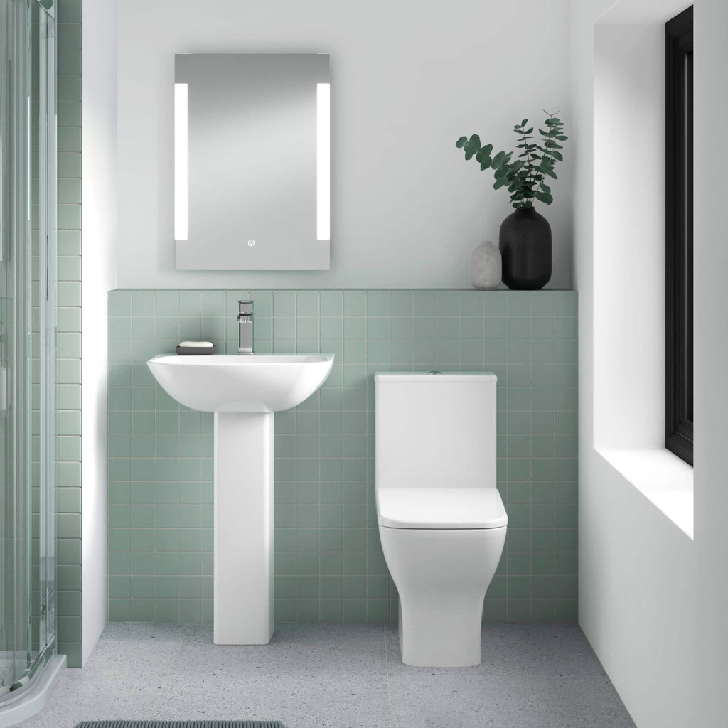 Modern minimalist bathroom with green tiled wall, white toilet and pedestal sink, black vase with greenery, and sleek mirror with LED lighting for contemporary bathroom design.