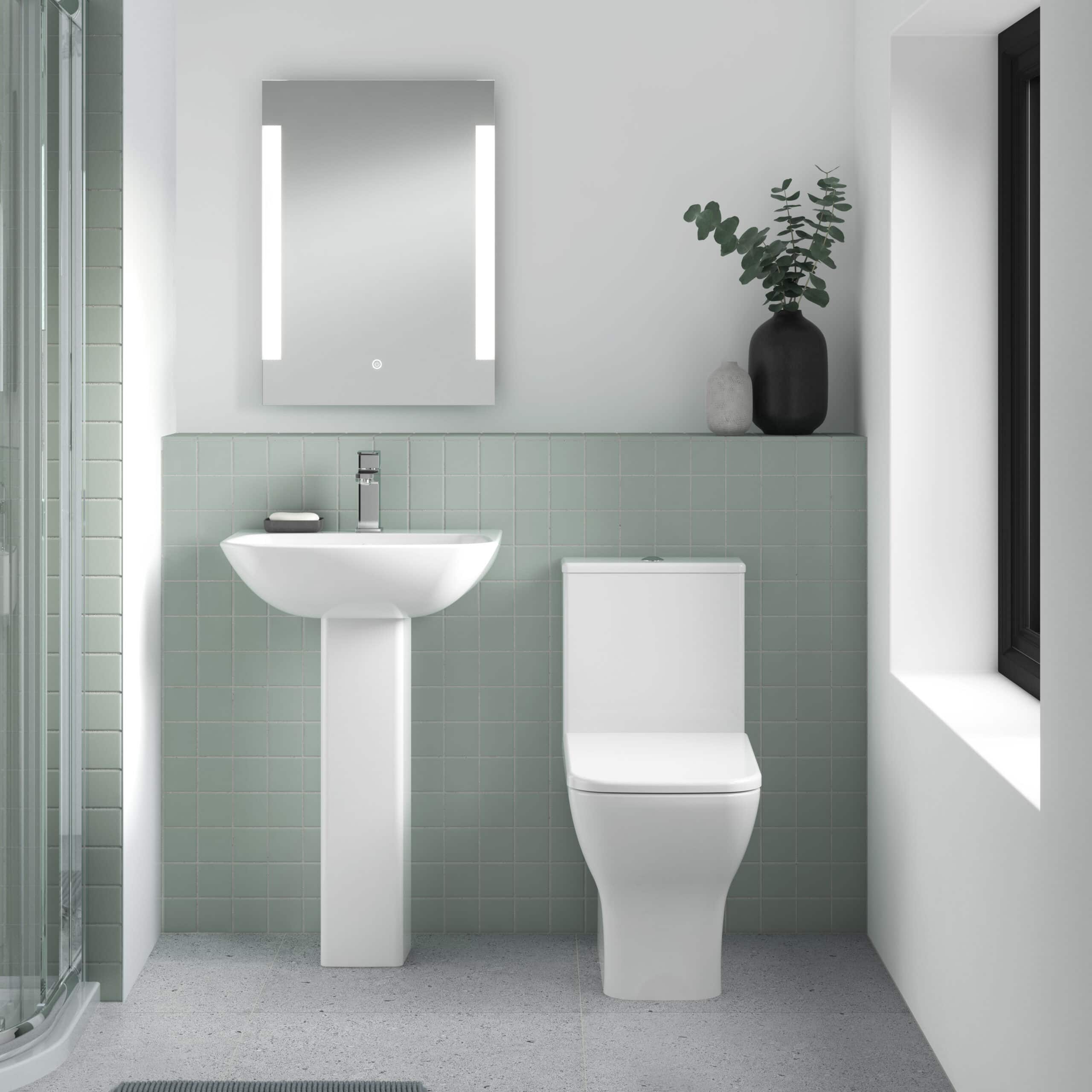 Modern minimalist bathroom with green tiled wall, white toilet and pedestal sink, black vase with greenery, and sleek mirror with LED lighting for contemporary bathroom design.