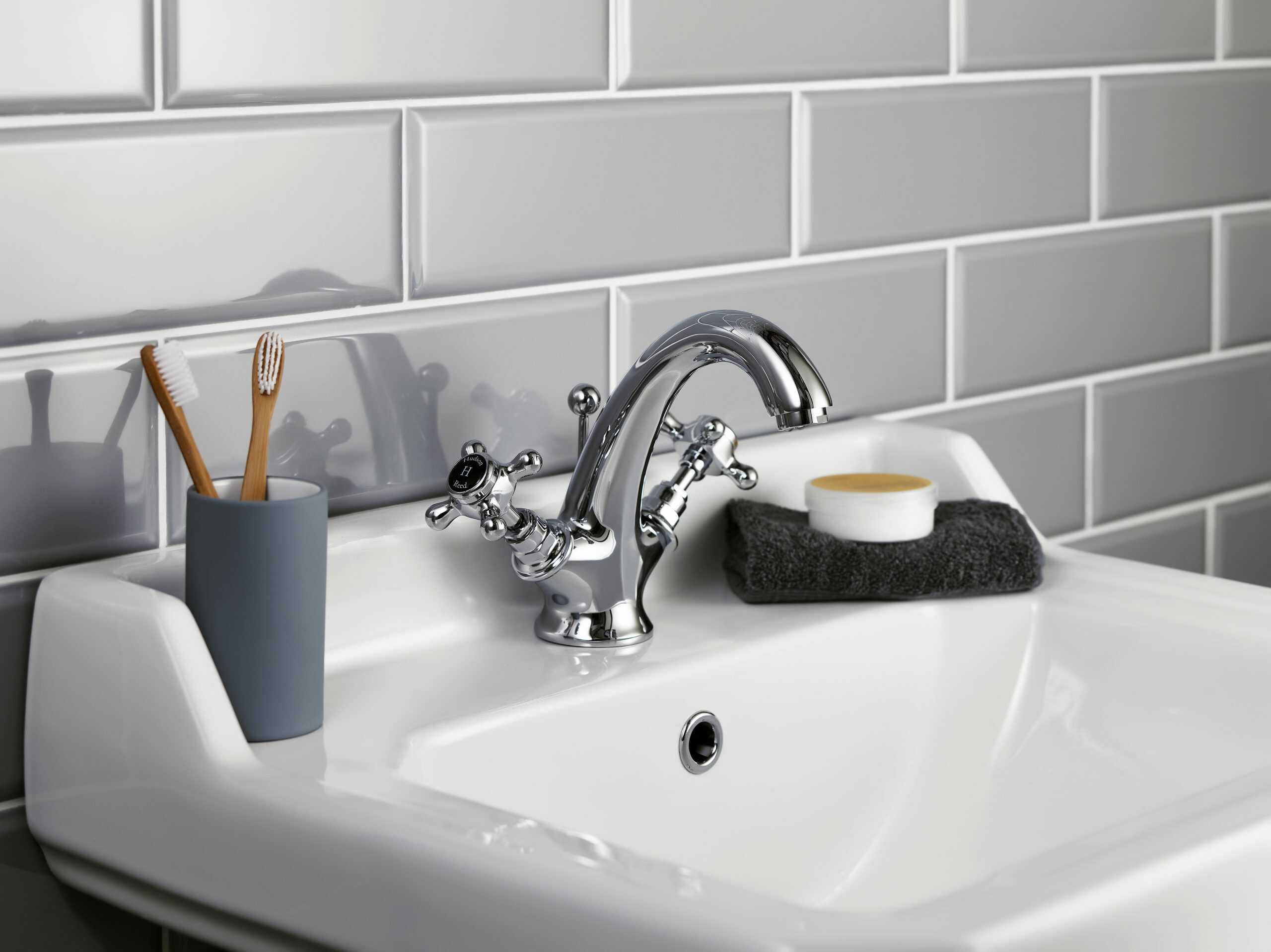 Polished chrome bathroom tap with classic cross handles on white ceramic sink, grey tiled wall backdrop, black washcloth, and toiletries for modern bathroom décor.