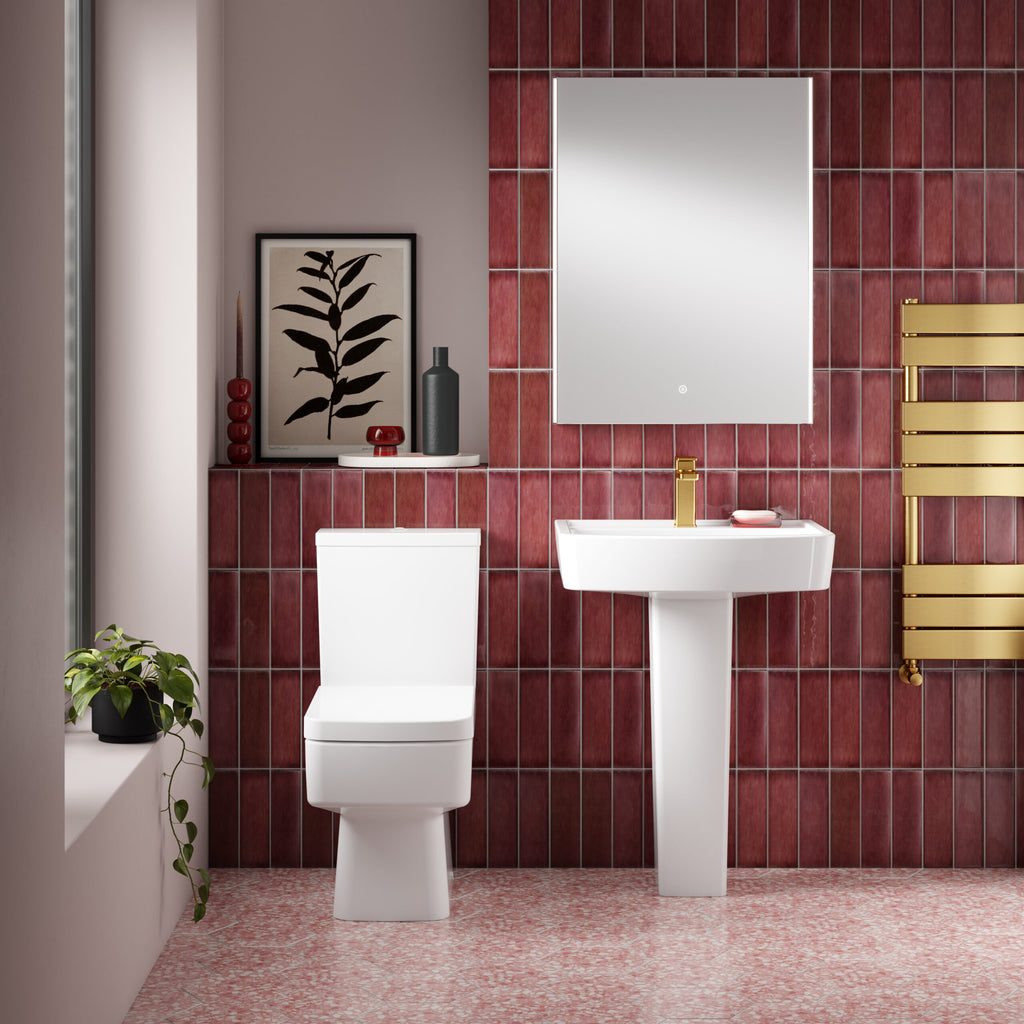 Modern bathroom with white toilet and pedestal sink, red tiled walls, gold heated towel rail, and decorative accessories, creating a stylish and functional space.