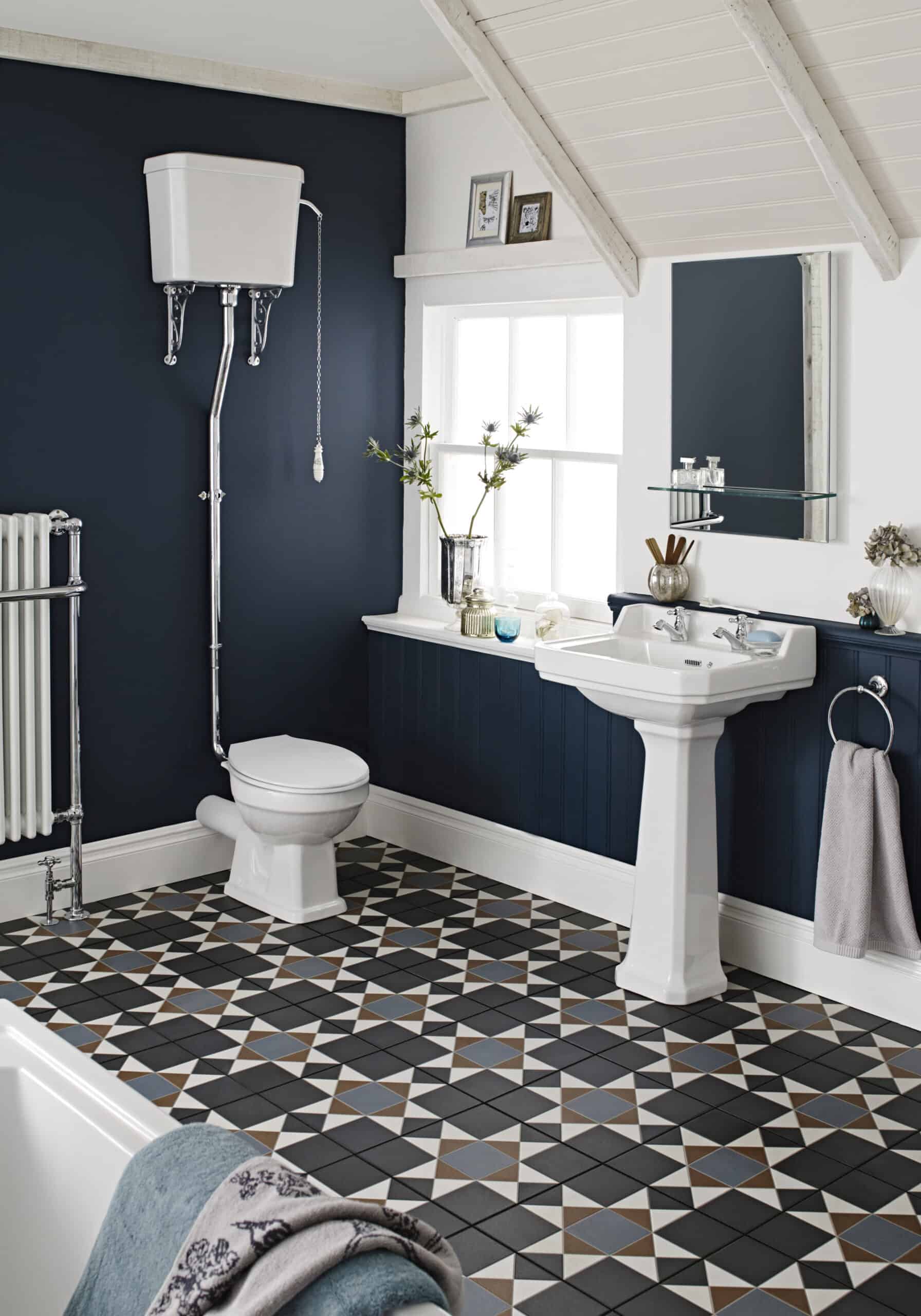 Toilet with high-level cistern and decorative plumbing in a stylish bathroom with vintage pedestal sink, patterned floor tiles, and navy wall paneling.