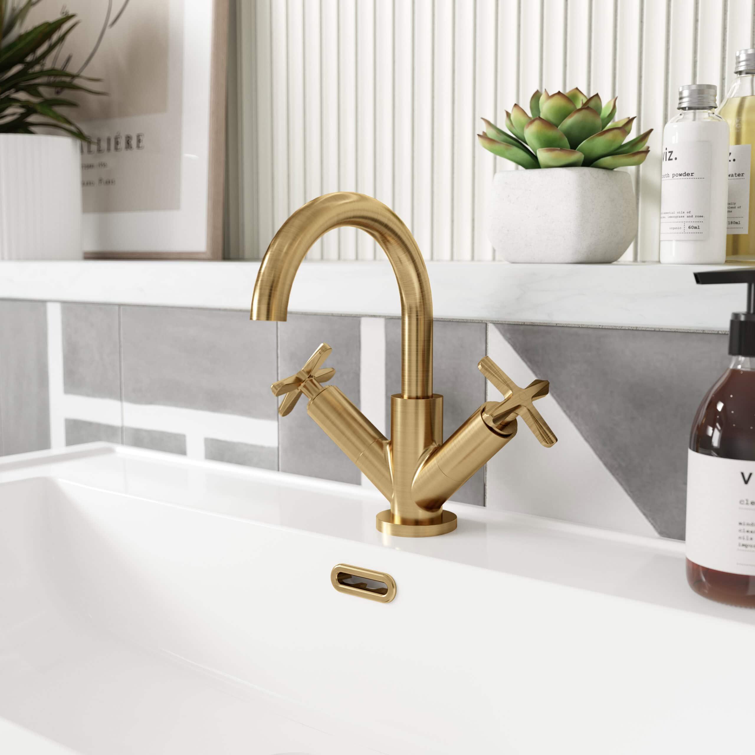 Sleek gold bathroom mixer tap with dual handles installed over a white sink in a modern bathroom setting. Botanical decorations and skincare products complement the stylish design.