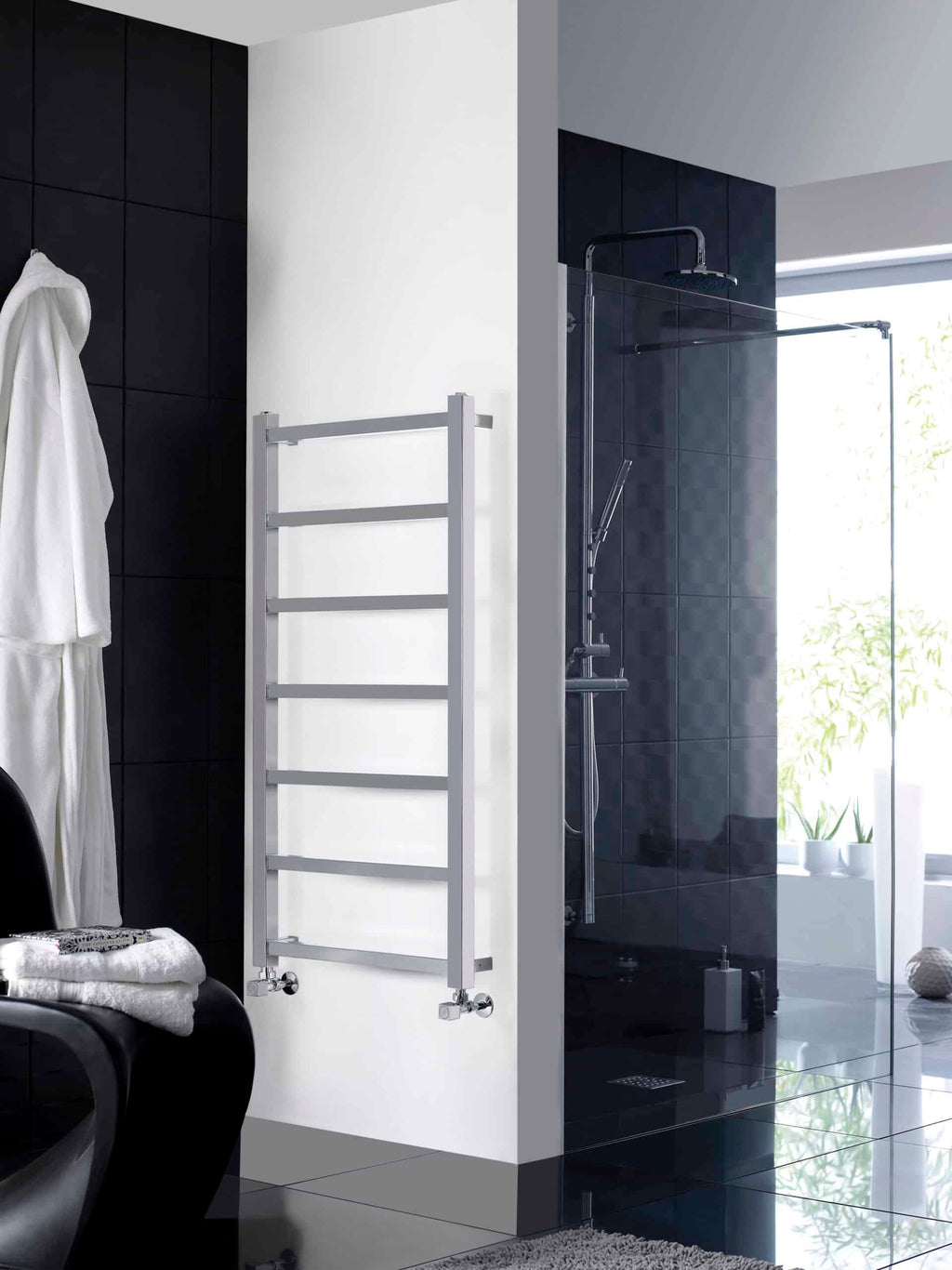 Modern white towel radiator in a contemporary bathroom setting, with a sleek black and white colour scheme, next to a stylish glass-enclosed shower and dark tiled walls, ideal for bathroom heating solutions.