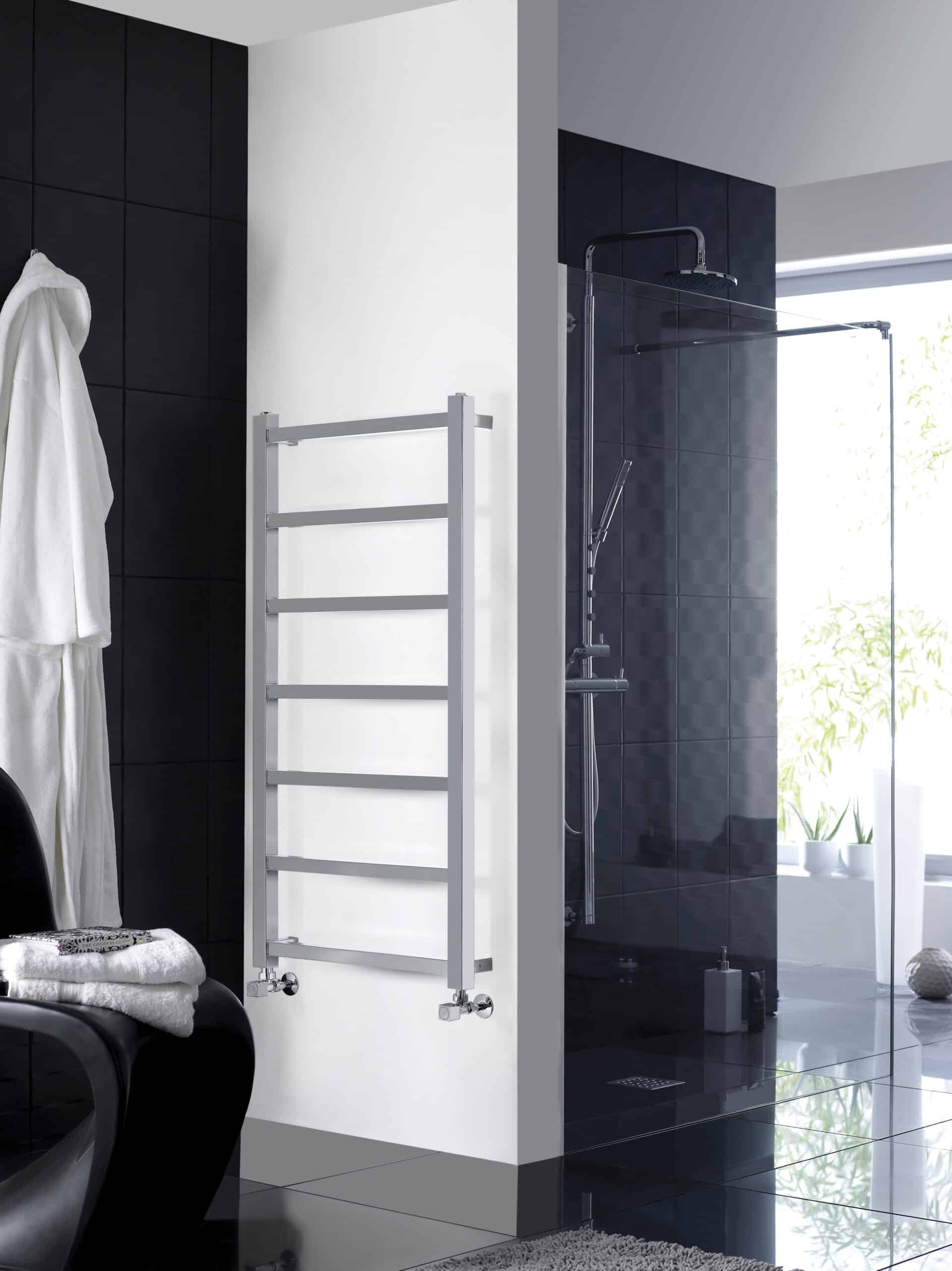 Modern white towel radiator in a contemporary bathroom setting, with a sleek black and white colour scheme, next to a stylish glass-enclosed shower and dark tiled walls, ideal for bathroom heating solutions.