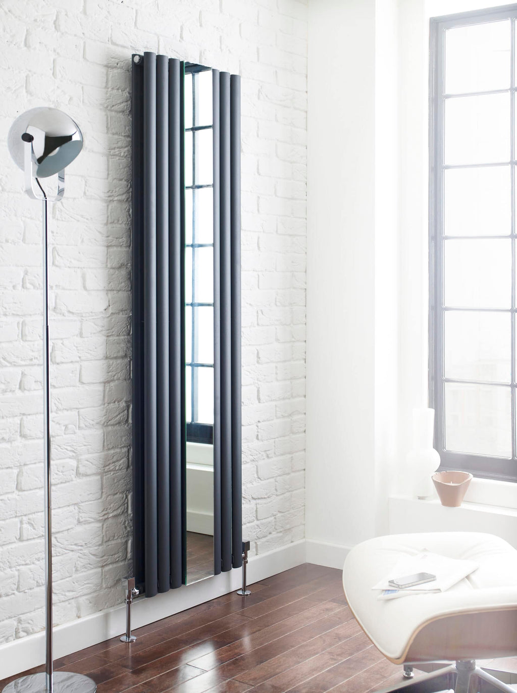 Sleek black vertical radiator with mirror in a modern bathroom setting, enhancing heating efficiency and style, next to a white textured brick wall and large window.