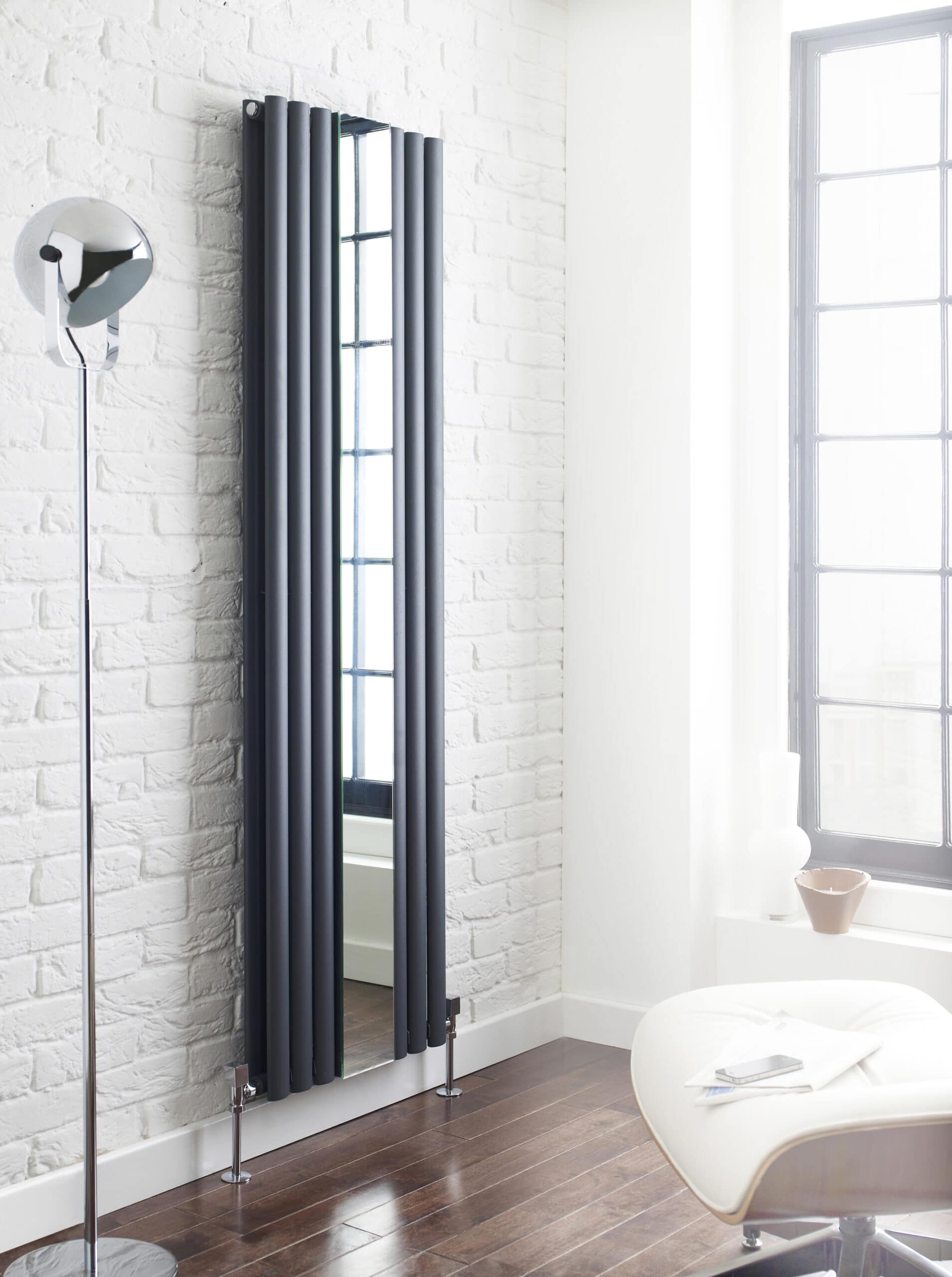 Sleek black vertical radiator with mirror in a modern bathroom setting, enhancing heating efficiency and style, next to a white textured brick wall and large window.