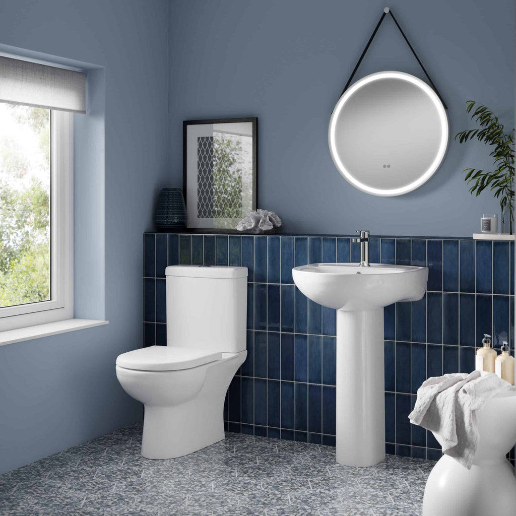 Sleek modern bathroom with white toilet, pedestal sink, round illuminated mirror, navy blue tiled wall, and decorative accessories, showcasing stylish and functional bathroom design.