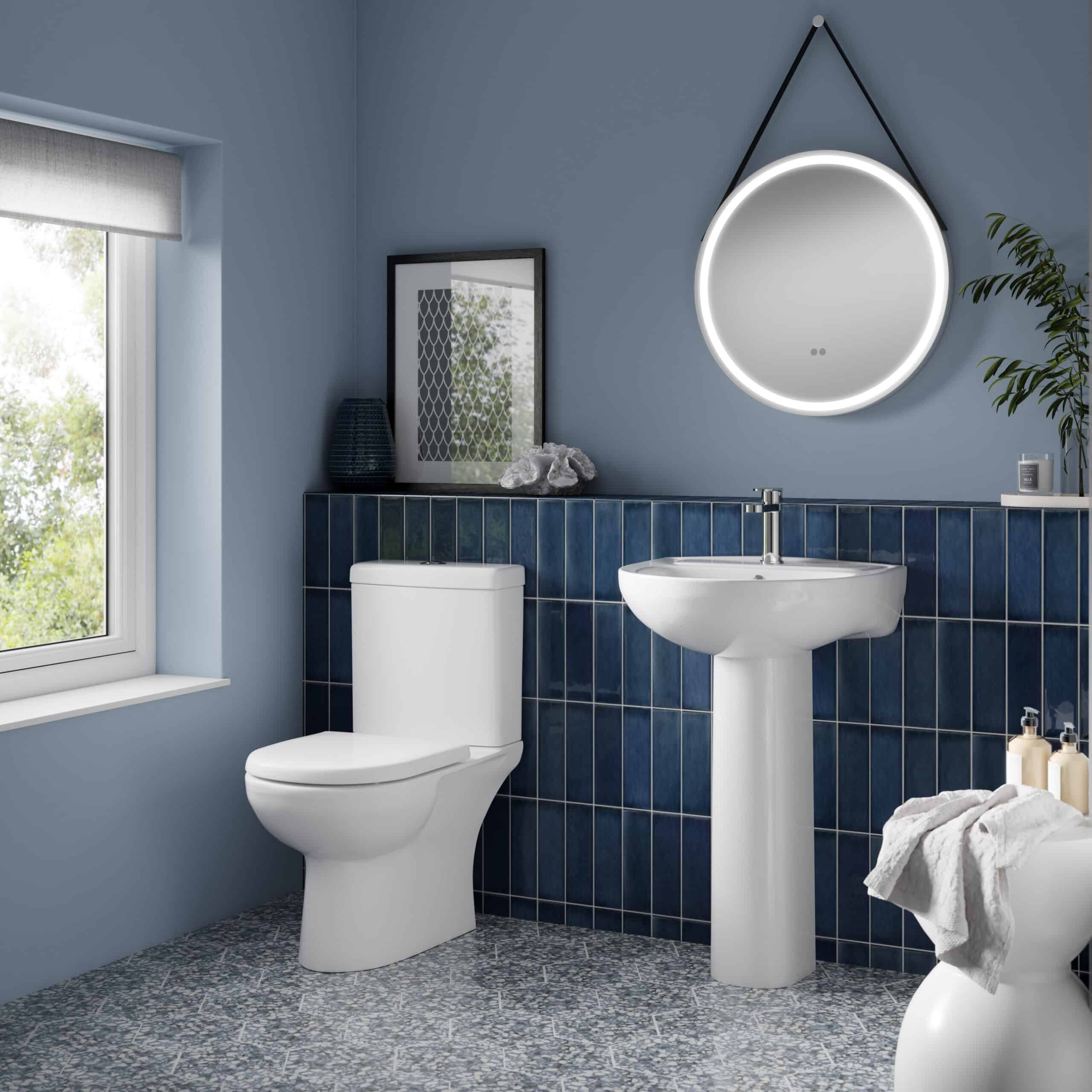 Sleek modern bathroom with white toilet, pedestal sink, round illuminated mirror, navy blue tiled wall, and decorative accessories, showcasing stylish and functional bathroom design.