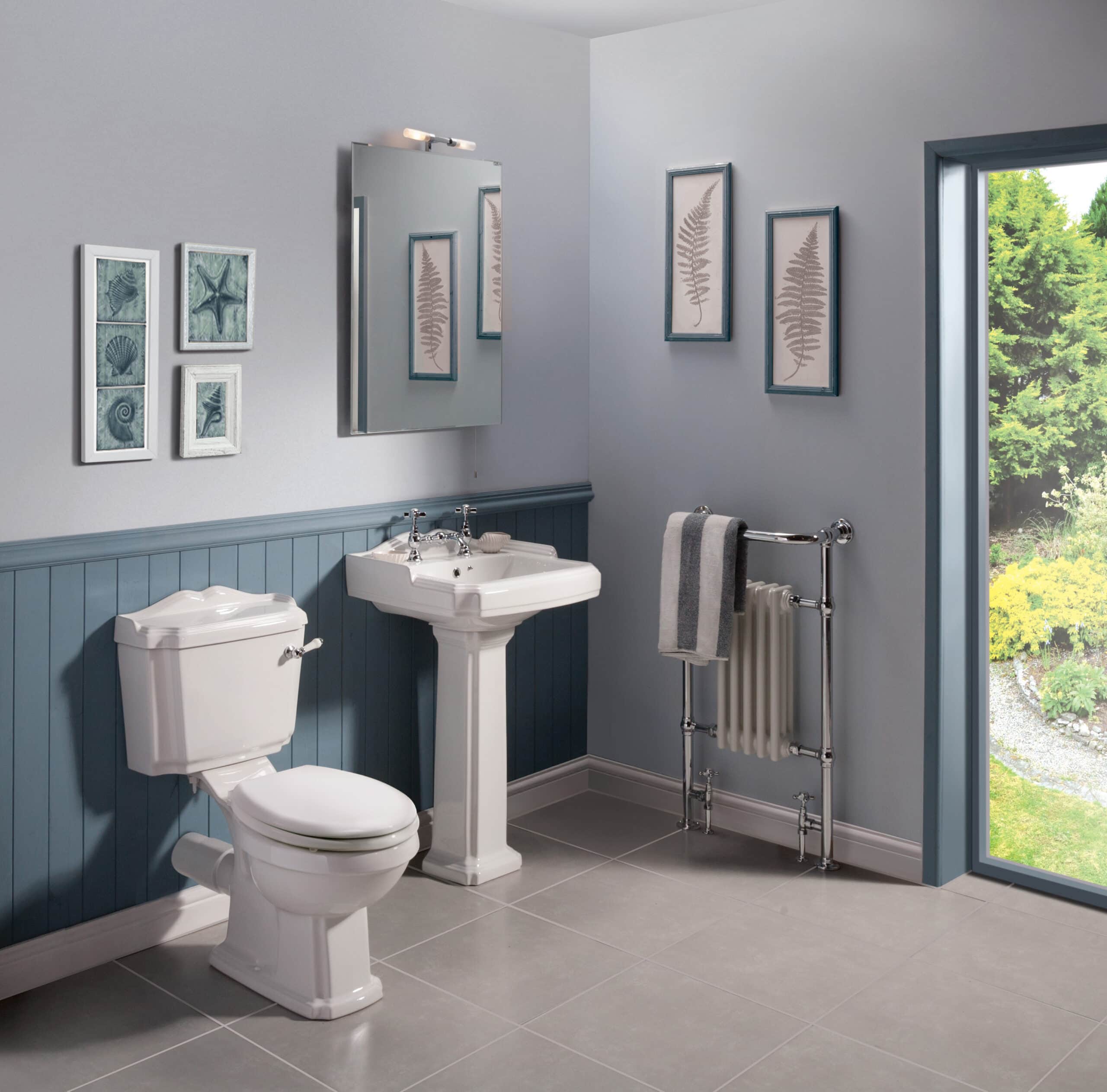 Elegant traditional bathroom with vintage white toilet and pedestal sink, framed coastal artwork, and a large window overlooking lush greenery, creating a serene bathroom space.