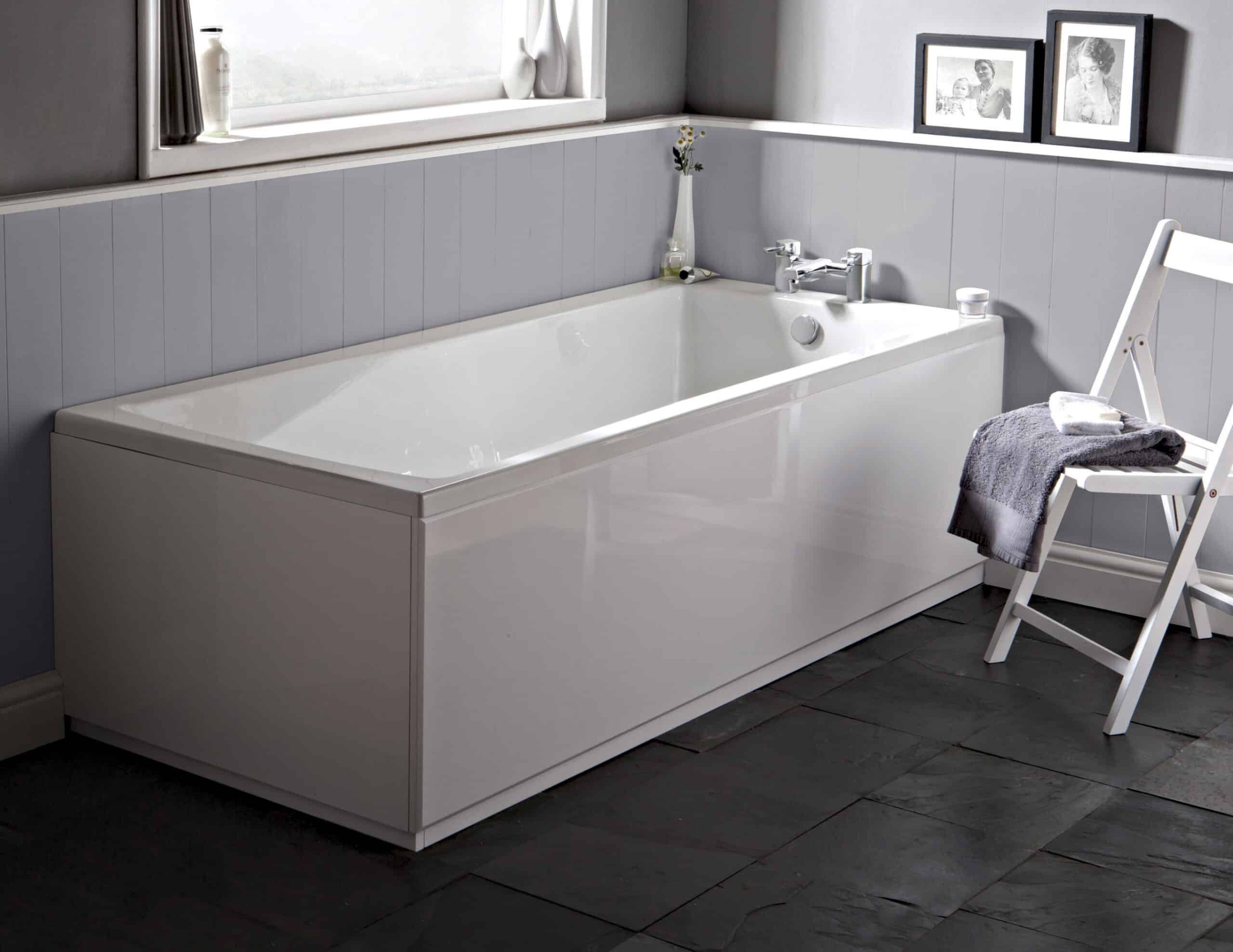 White modern bathtub in a contemporary bathroom setting with grey wall cladding and black slate flooring, featuring a white wooden chair with towels, and black-and-white framed photos on the wall.