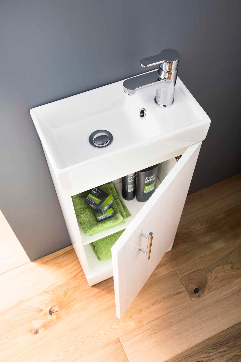 Contemporary white bathroom sink with chrome faucet, open cabinet storage with green towels and skincare products, sleek modern design, ideal for modern bathroom interiors.
