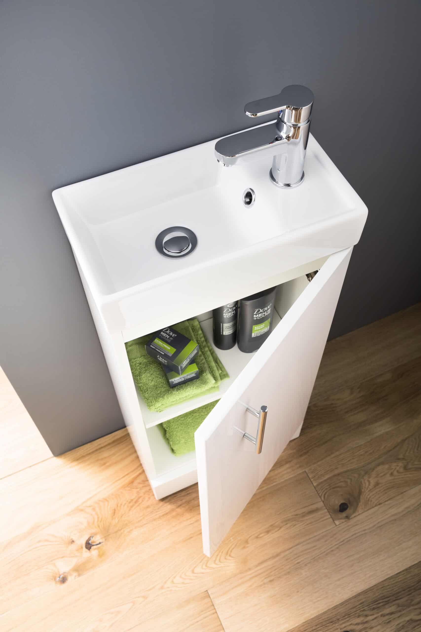 Contemporary white bathroom sink with chrome faucet, open cabinet storage with green towels and skincare products, sleek modern design, ideal for modern bathroom interiors.