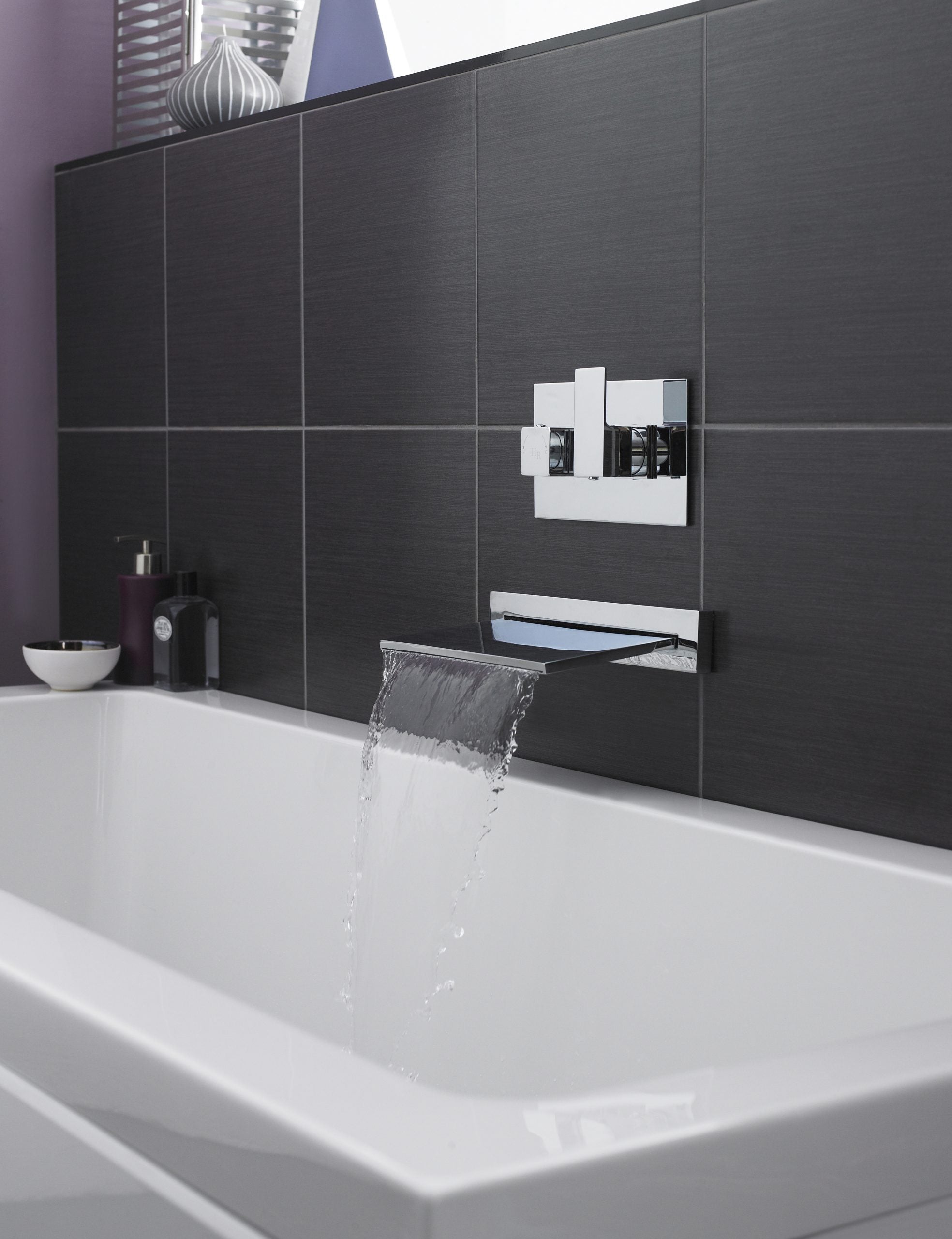 Sleek modern bathtub with waterfall faucet and dark grey tiled wall, showcasing contemporary bathroom fixtures and design.