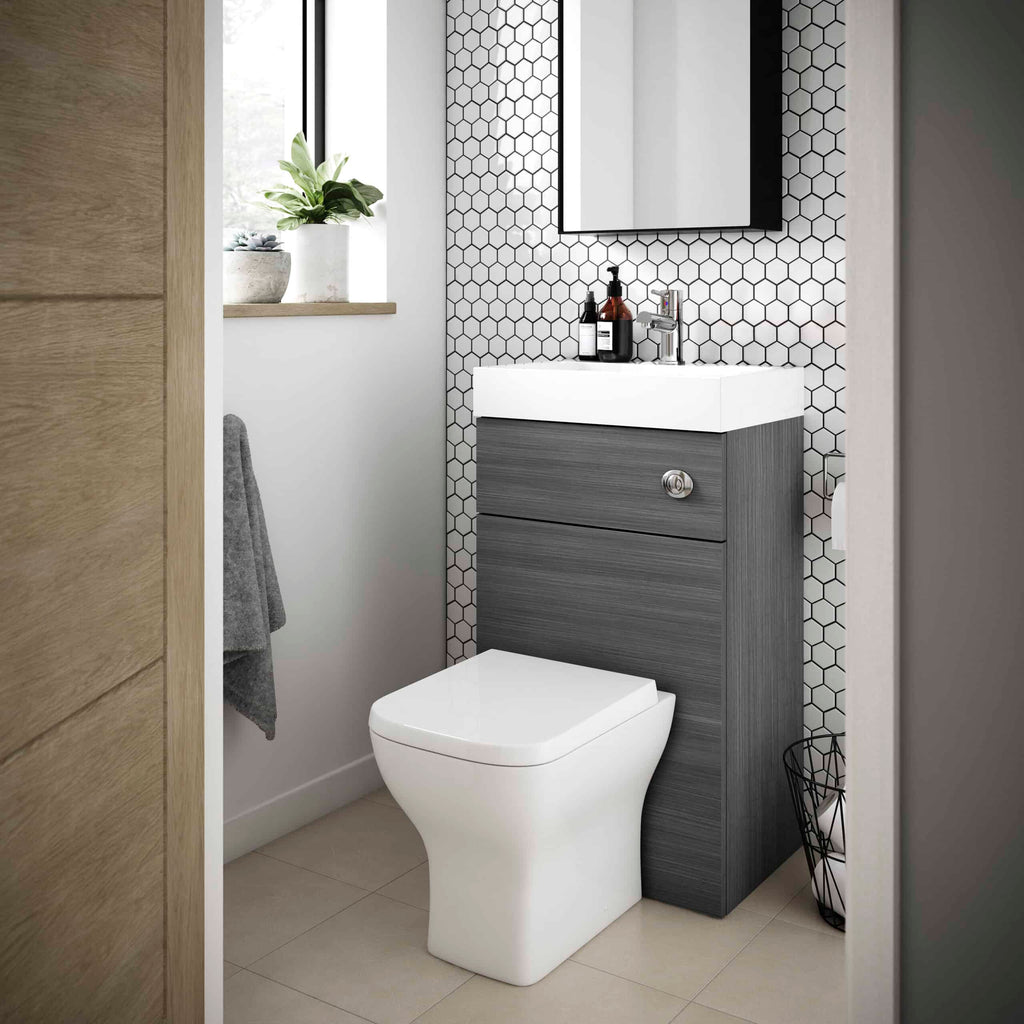 Sleek modern bathroom with a wall-mounted vanity with grey wood finish, white ceramic sink, black hex tile wall, small potted plant on windowsill, grey towel on side, and white toilet, minimalist design.