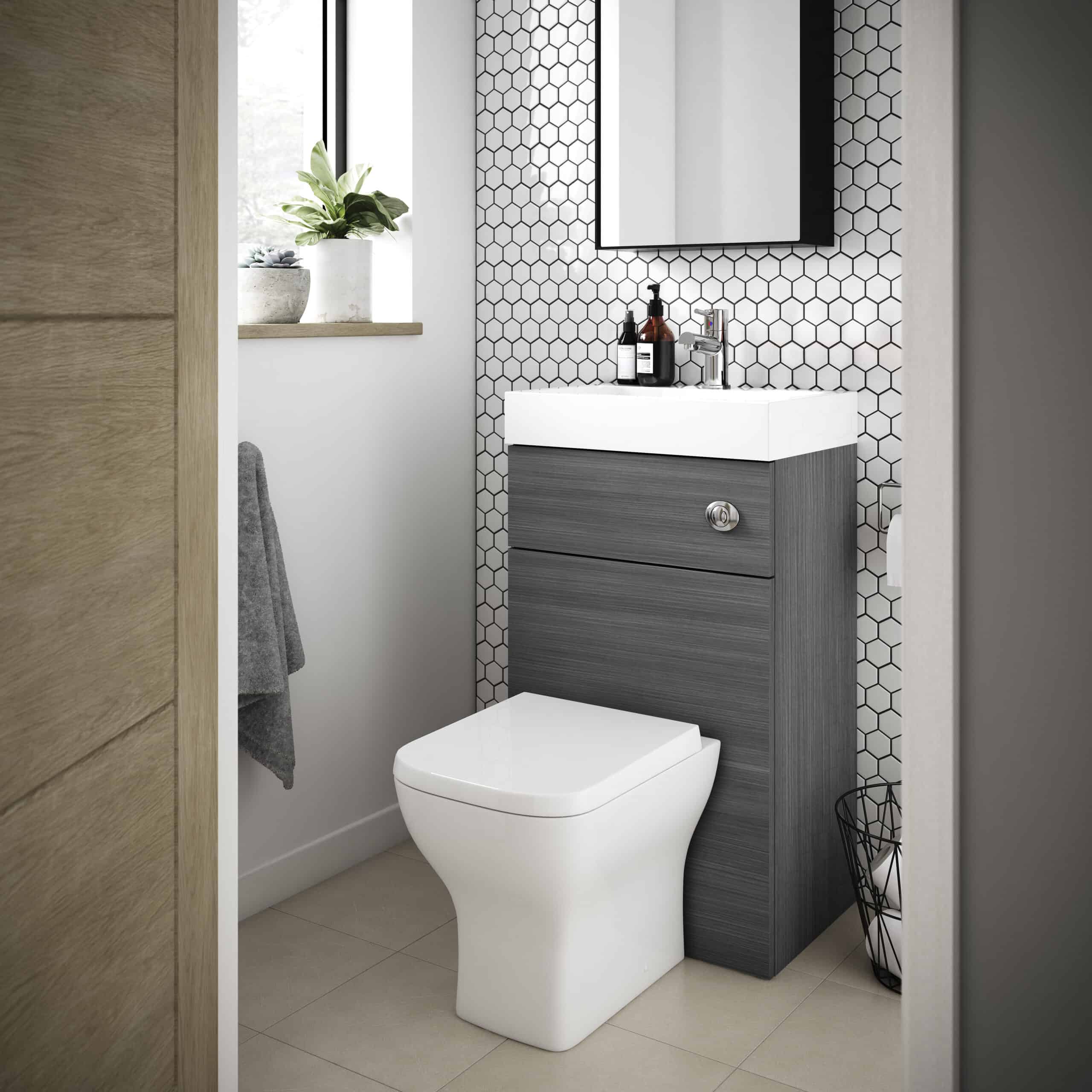 Sleek modern bathroom with a wall-mounted vanity with grey wood finish, white ceramic sink, black hex tile wall, small potted plant on windowsill, grey towel on side, and white toilet, minimalist design.