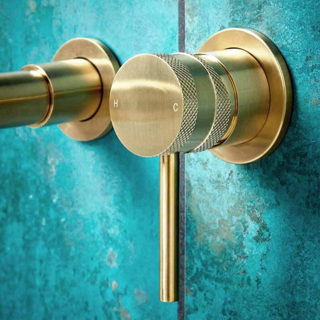 Shiny brass thermostatic shower control valve with textured grip, mounted on a turquoise tiled wall, representing high-quality bathroom fixtures for modern bathrooms.