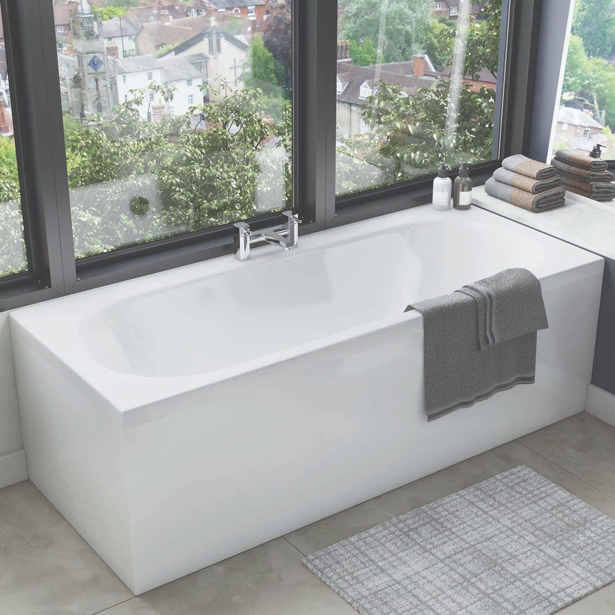 Sleek white freestanding bathtub with modern chrome tap, positioned by large floor-to-ceiling windows with a cityscape view, complemented by grey towels and bathroom essentials, in a contemporary bathroom setting.
